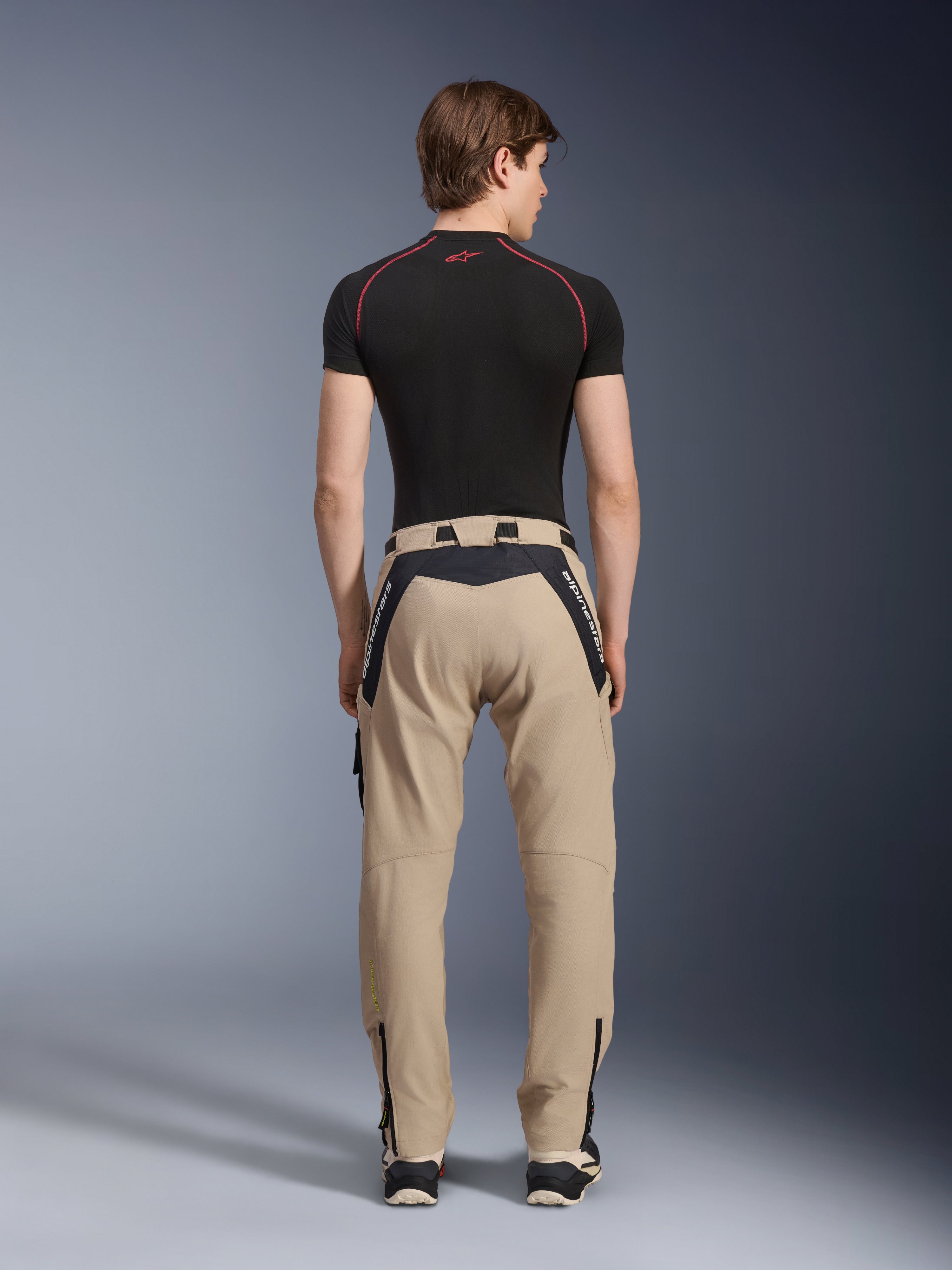 Alpinestars Acteon Hose in Sand, moderne urbane Motorradhose, Rückansicht, schwarze Einsätze mit weißem Alpinestars-Branding, schmal zulaufende Passform mit Reißverschlüssen am Saum, getragen mit schwarzem kurzärmeligem Baselayer und Sneakern.