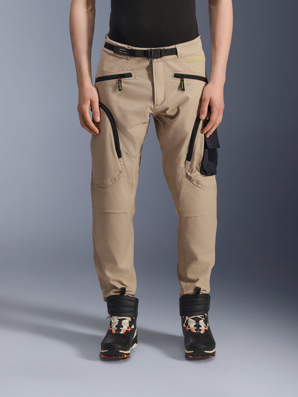 Alpinestars Acteon Hose, urbane Motorradhose, Sandbeige, Cargo-Design mit mehreren Taschen, schwarze umgekehrte Spiralreißverschlüsse, integrierter Gürtel und schmal zulaufende Passform, getragen mit schwarz-orangen Stiefeln