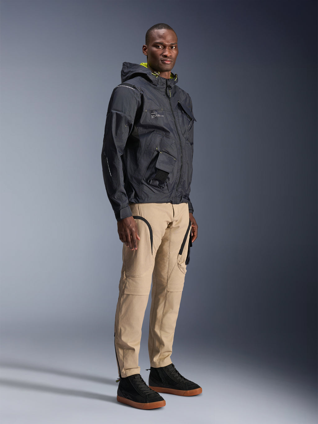 Alpinestars Acteon Hose, urbane Motorradhose, Farbe Sand, getragen von einem Model mit einer dunklen Funktionsjacke und schwarzen Sneakern, mit schmal zulaufender Passform, schwarzen Reißverschluss-Akzenten und Cargotaschen, zeitgemäßes, vom Motorsport inspiriertes Design
