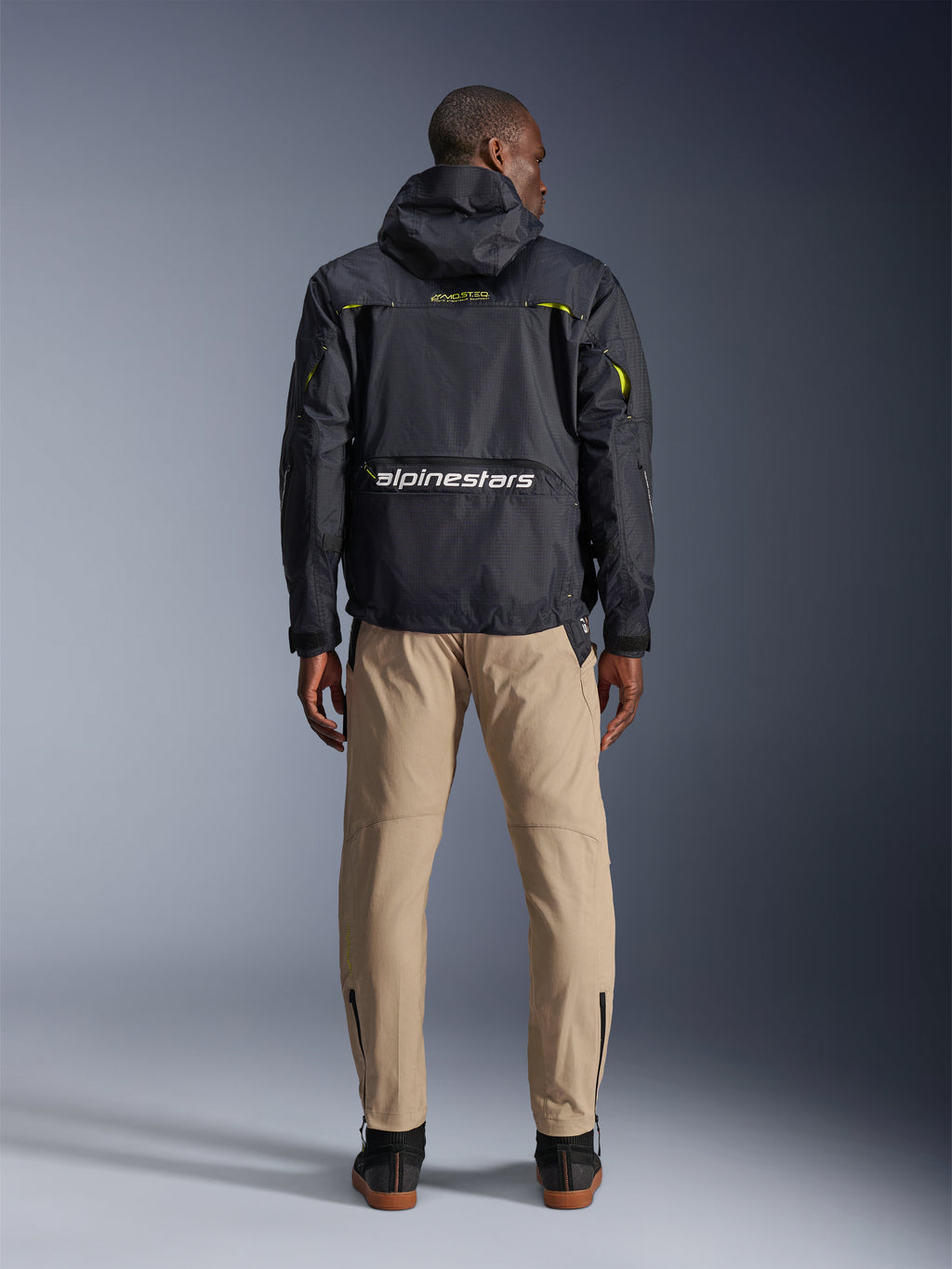 Alpinestars Acteon Hose, Urbane Motorradhose, Sand, Rückansicht an einem Model mit schwarzer technischer Kapuzenjacke und dunklen Stiefeln, schmal zulaufendes Bein mit Knöchel-Reißverschlüssen, moderne Silhouette