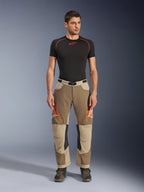 Alpinestars Maxdura Dual Hose, Adventure- und Dual-Sport-Motorradhose, Walnut Aluminum, getragen mit einem schwarzen kurzärmeligen Kompressionsshirt und Adventure-Stiefeln, mit orangefarbenen Belüftungsreißverschlüssen und roten Logos auf den Oberschenkeln, schwarzer Taillengürtel mit Schnalle, Über-den-Stiefel-Design zum Motorradfahren.