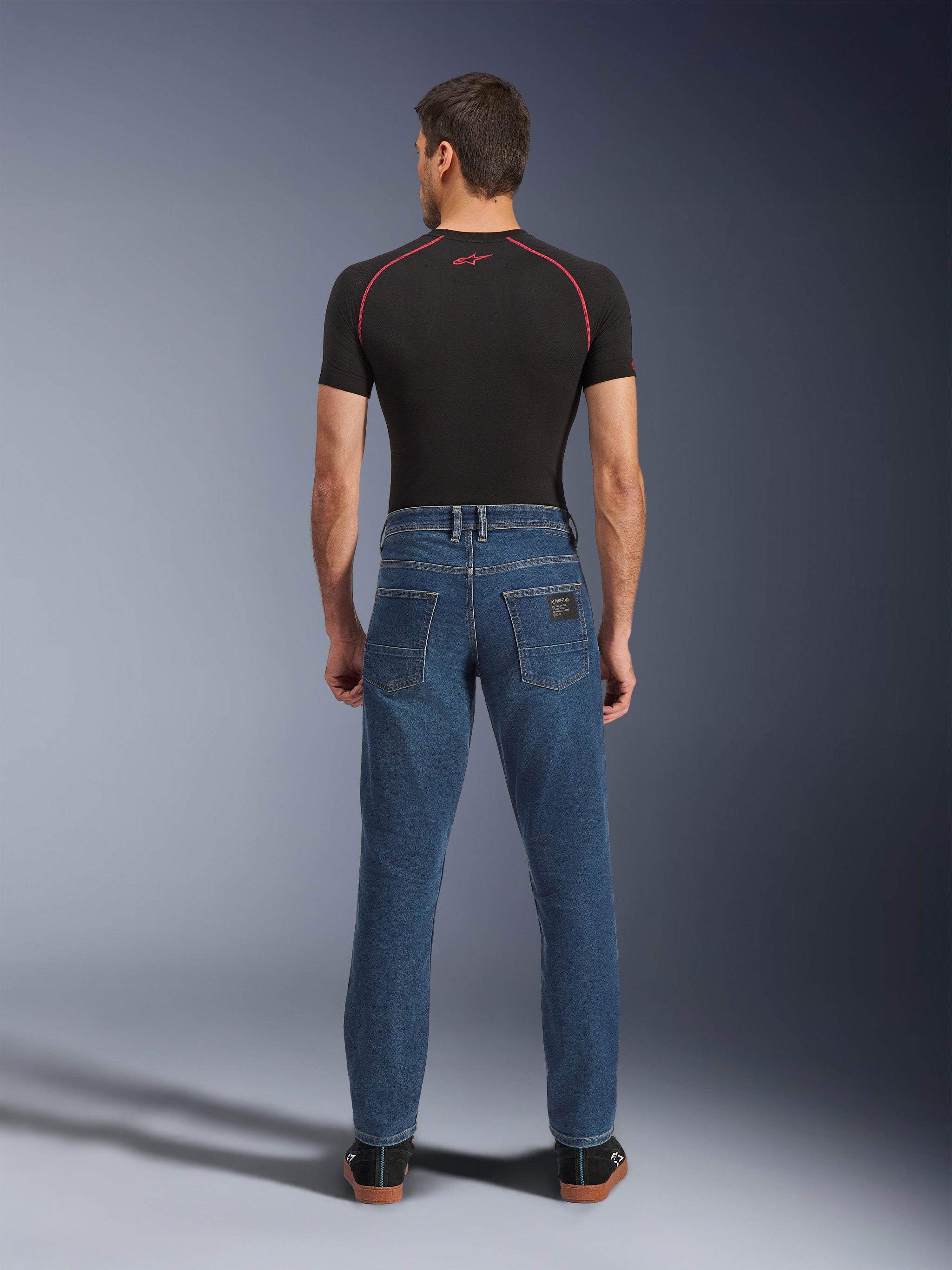 Alpinestars Flex-AST 5 Denim-Hose, Indigo Washed Blau, Rückansicht mit konisch zulaufender urbaner Passform, getragen von einem Modell mit schwarzem Performance-T-Shirt und Sneakern, lässige Motorrad-Schutzkleidung