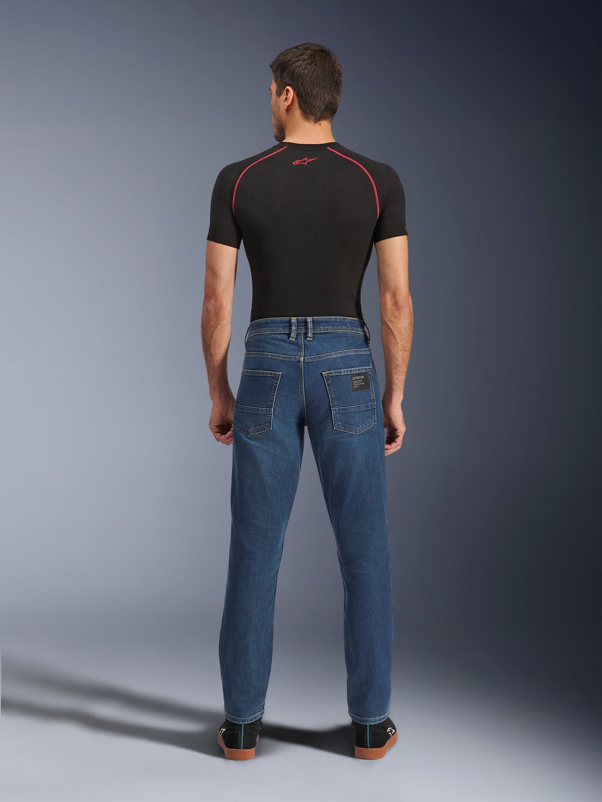 Mann in der Rückansicht trägt Alpinestars Flex-AST 5 Denim-Hose in Indigo Washed, urbane Motorradjeans mit moderner konischer Passform und Gesäßtaschen, kombiniert mit einem schwarzen kurzärmeligen Baselayer und lässigen schwarzen Schuhen.