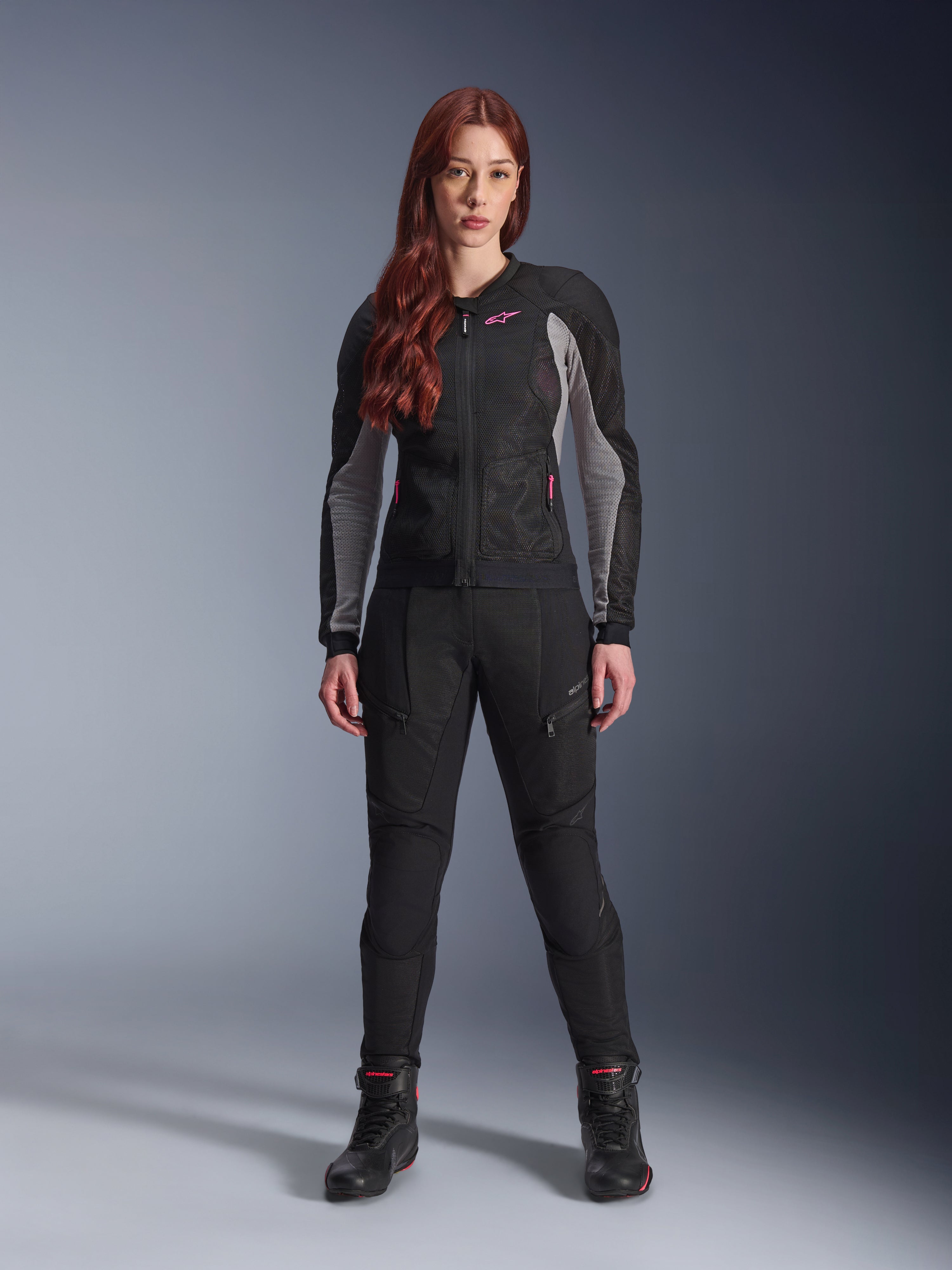 Alpinestars Stella Troop-Air Jacket and Pants, Women's Motorcycle Riding Gear, Black with Ash Gray and Pink Fluo accents, worn by a female model with red hair, featuring mesh ventilation and protective design, studio standing pose.