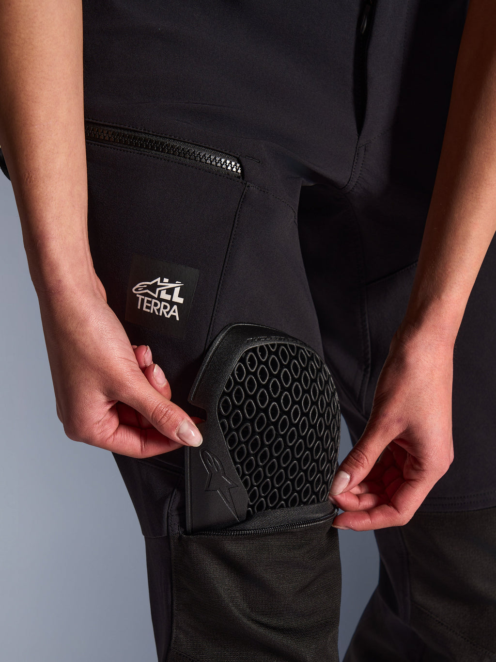 Nahaufnahme der Alpinestars Stella Flex-AST Explorer Hose in Schwarz, zeigt die externe Protektorentasche beim Einsetzen des Nucleon Flex Plus Knieprotektors, Detail der Wabenstruktur, „ALL TERRA“ Branding auf dem Oberschenkel, funktionale Adventure-Textilausrüstung für Damen