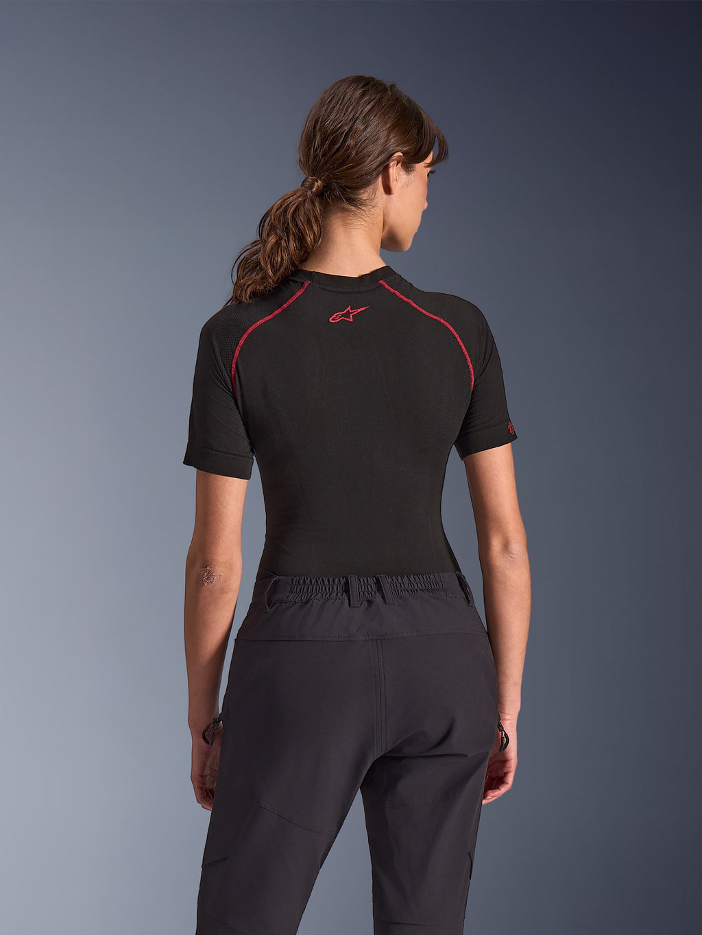 Frau trägt Alpinestars Stella Flex-AST Explorer Hose, schwarze Adventure-Motorradhose, Rückansicht, kombiniert mit einem schwarzen kurzärmeligen Oberteil mit roten Nähten, betont die atmungsaktive Textilkonstruktion und den elastischen Hosenbund.
