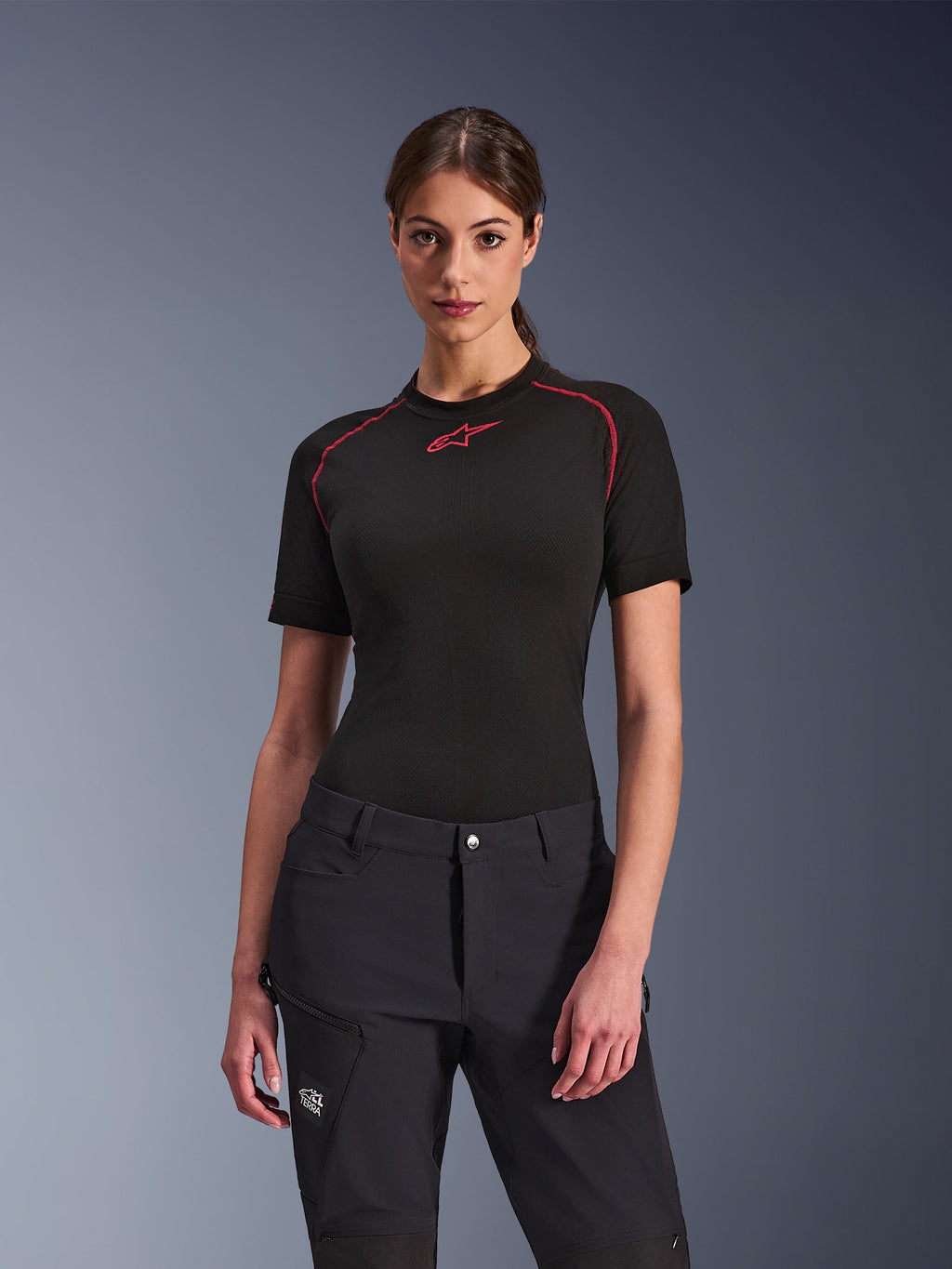 Frau in Alpinestars Stella Flex-AST Explorer Hose in Schwarz, kombiniert mit einem schwarzen kurzärmeligen Base Layer mit roten Akzenten, Adventure-Fahrbekleidung mit Cargo-Taschen und All Terra-Branding