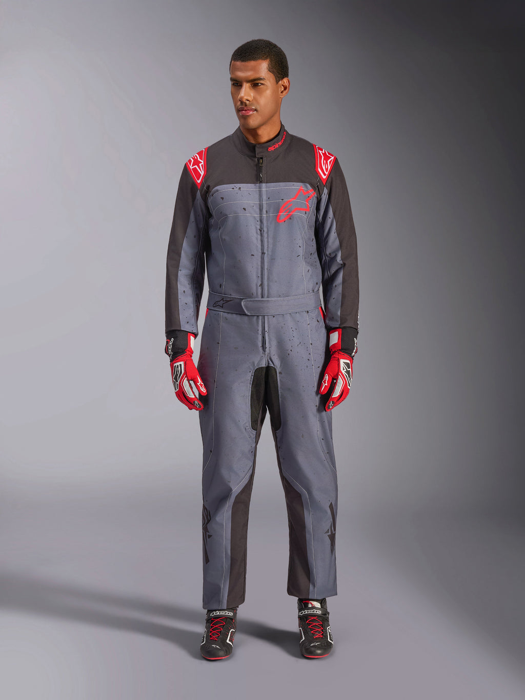 Alpinestars KMX-9 V3 Graphic 6 Anzug, Kart-Anzug, Anthrazit-Schwarz und Rot, grau gesprenkeltes Grafikmuster, schwarze Einsätze an den Innenseiten von Armen und Beinen, rote Alpinestars-Logos auf Schultern und Brust, getragen mit roten Rennhandschuhen und schwarzen Schuhen