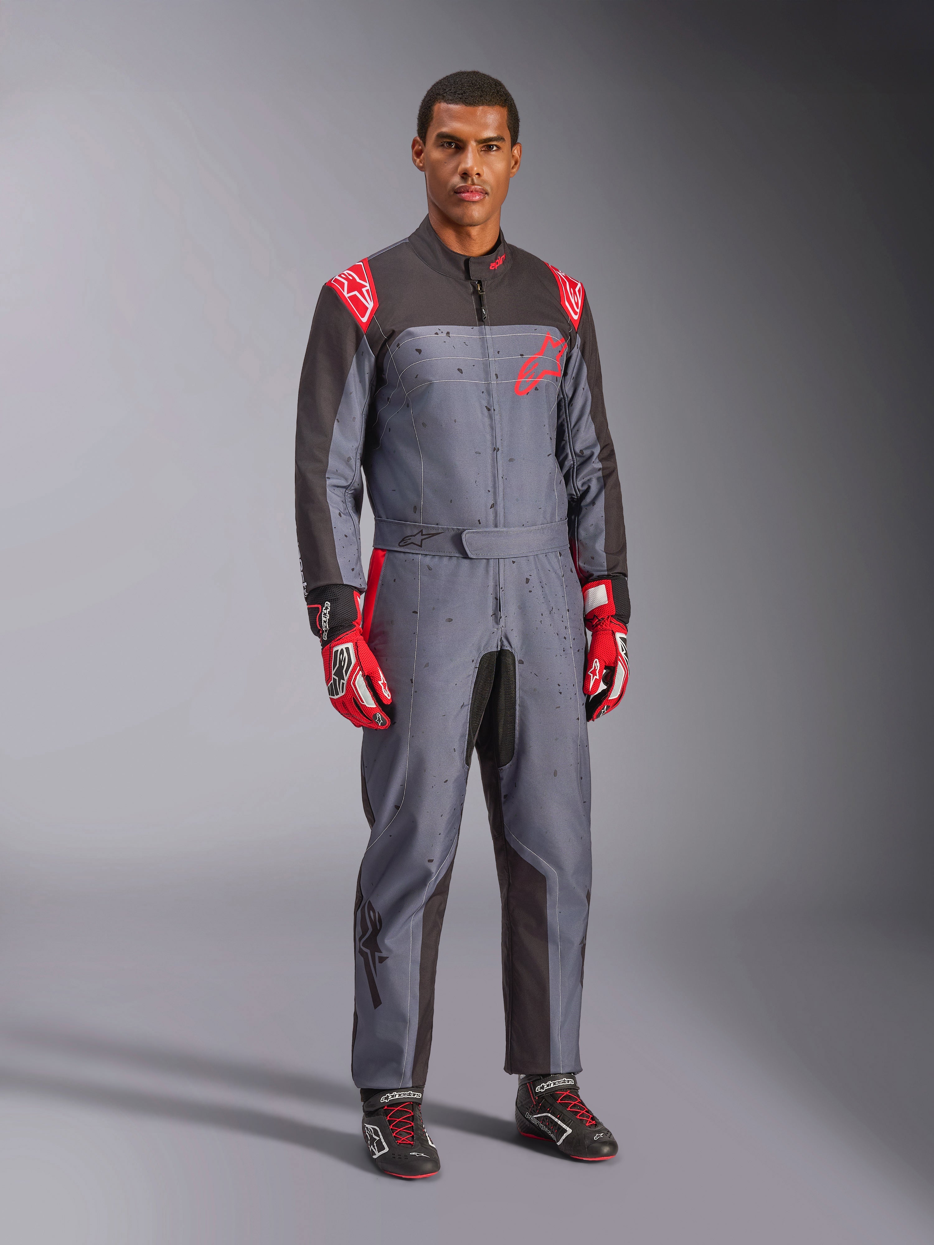 Alpinestars KMX-9 V3 Graphic 6 Karting-Anzug, Grau und Anthrazit mit roten Akzenten, mit einer einzigartigen Splash-Grafik auf der Brust und den Beinen, kombiniert mit roten Rennhandschuhen und schwarzen Karting-Schuhen, Ganzkörper-Studioaufnahme