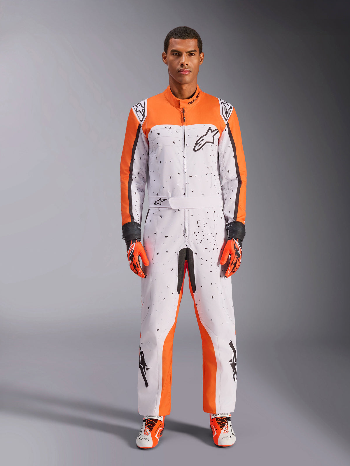 Alpinestars KMX-9 V3 Graphic 6 Suit, Karting Suit, White Orange and Black, featuring a black speckled pattern, worn with matching orange and black racing gloves and shoes, one-piece motorsport protective gear with bold logos and athletic design.