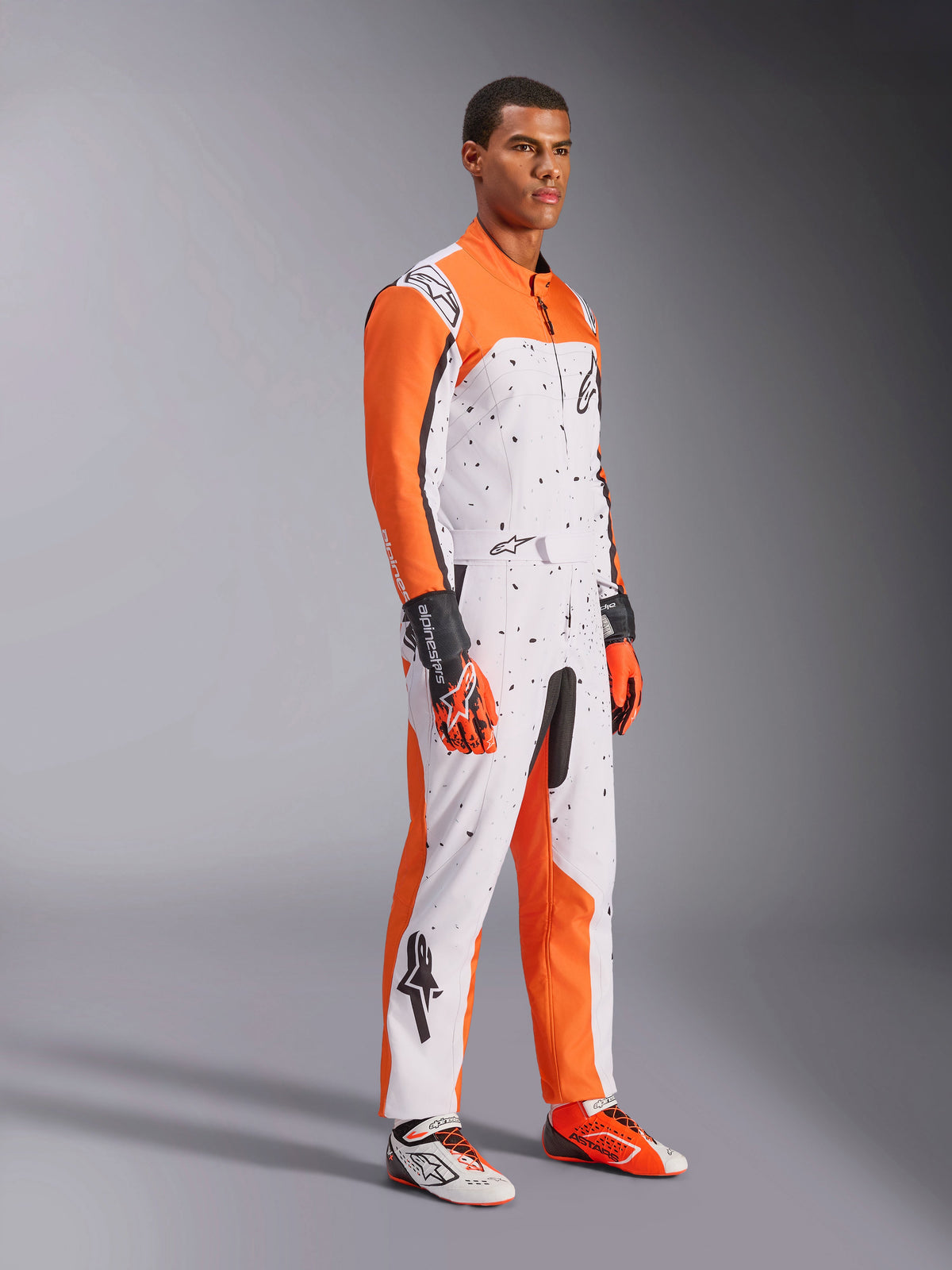 Alpinestars KMX-9 V3 Graphic 6 Kart-Anzug, Weiß, Orange und Schwarz, mit gesprenkeltem Muster und leuchtend orangefarbenen Ärmeln, getragen von einem Modell mit passenden Rennhandschuhen und Schuhen, professionelle zweilagige Motorsport-Ausrüstung.