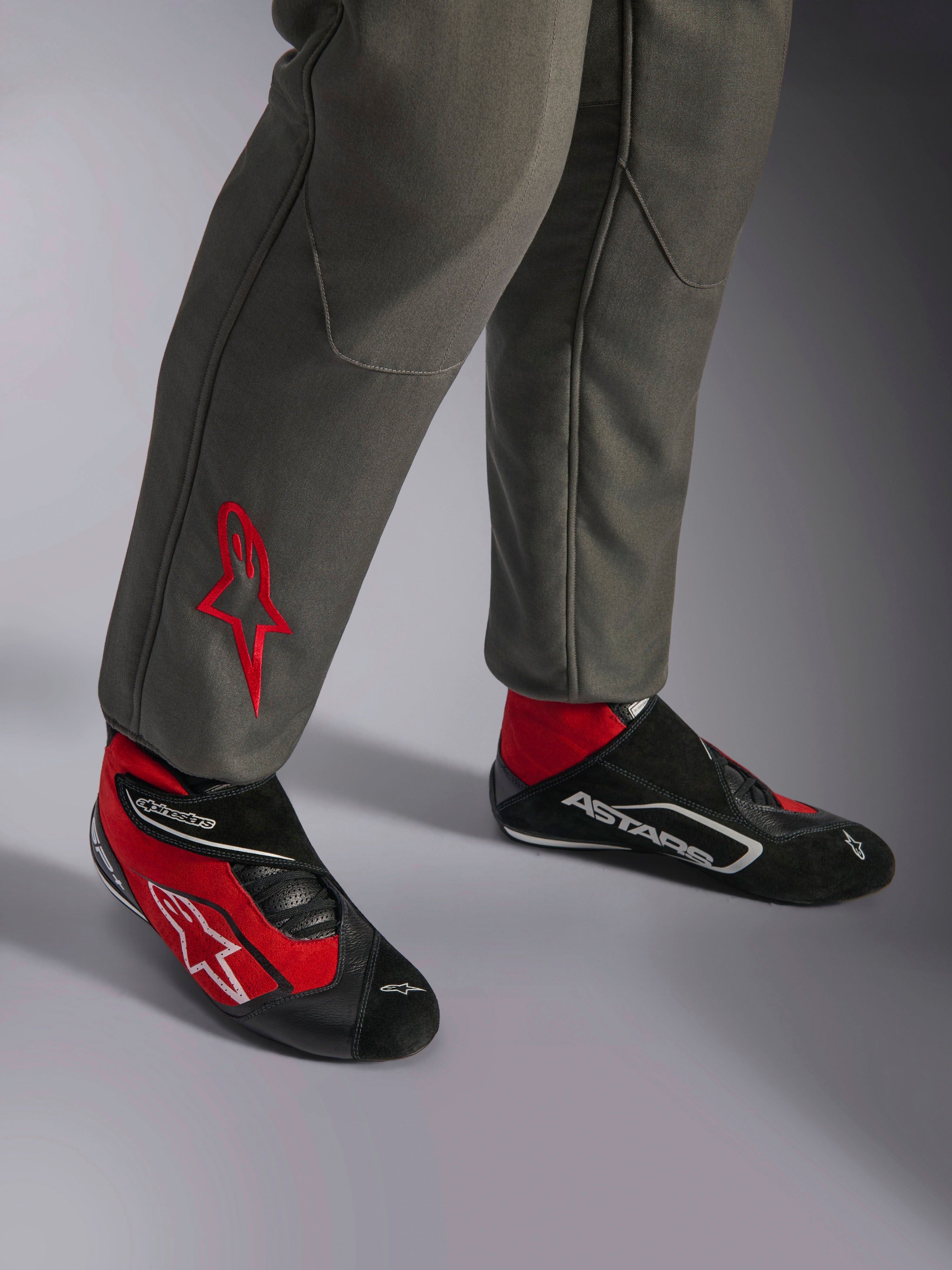Alpinestars GP Race V3 Anzug FIA, Rennanzug in Anthrazit-Rot, Nahaufnahme der Unterschenkel mit besticktem rotem Stern-Logo, kombiniert mit rot-schwarzen Rennstiefeln, professionelle Automobil-Ausrüstung für Fahrerschutz und Komfort