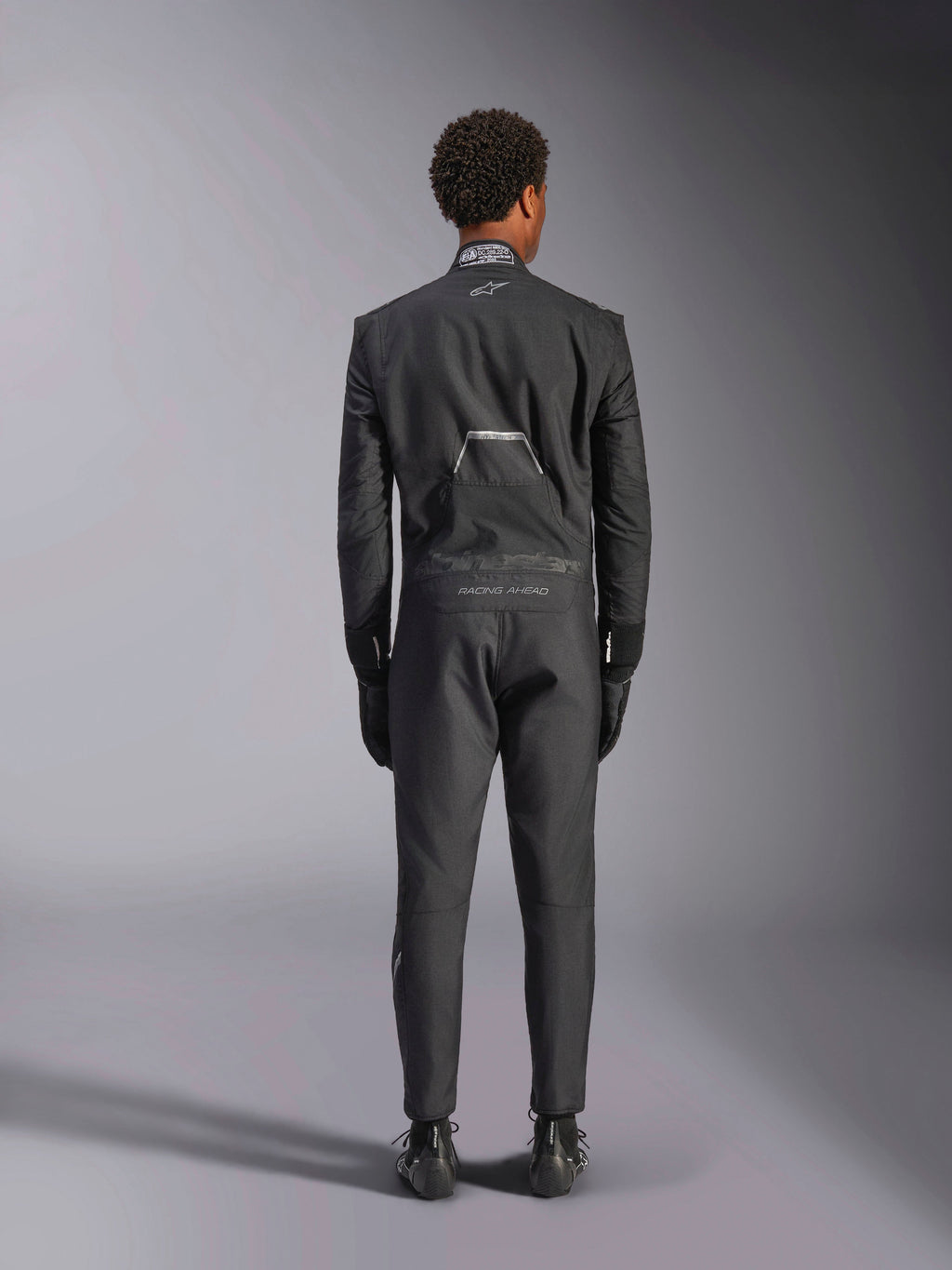 Alpinestars Hypertech V3 Anzug, Schwarz Dark Shadow, Rückansicht eines professionellen einteiligen Auto-Rennanzugs, mit FIA-Zertifizierungsetikett am Kragen und „Alpinestars“- sowie „RACING AHEAD“-Branding am unteren Rücken, getragen mit passenden schwarzen Rennhandschuhen und Stiefeln.