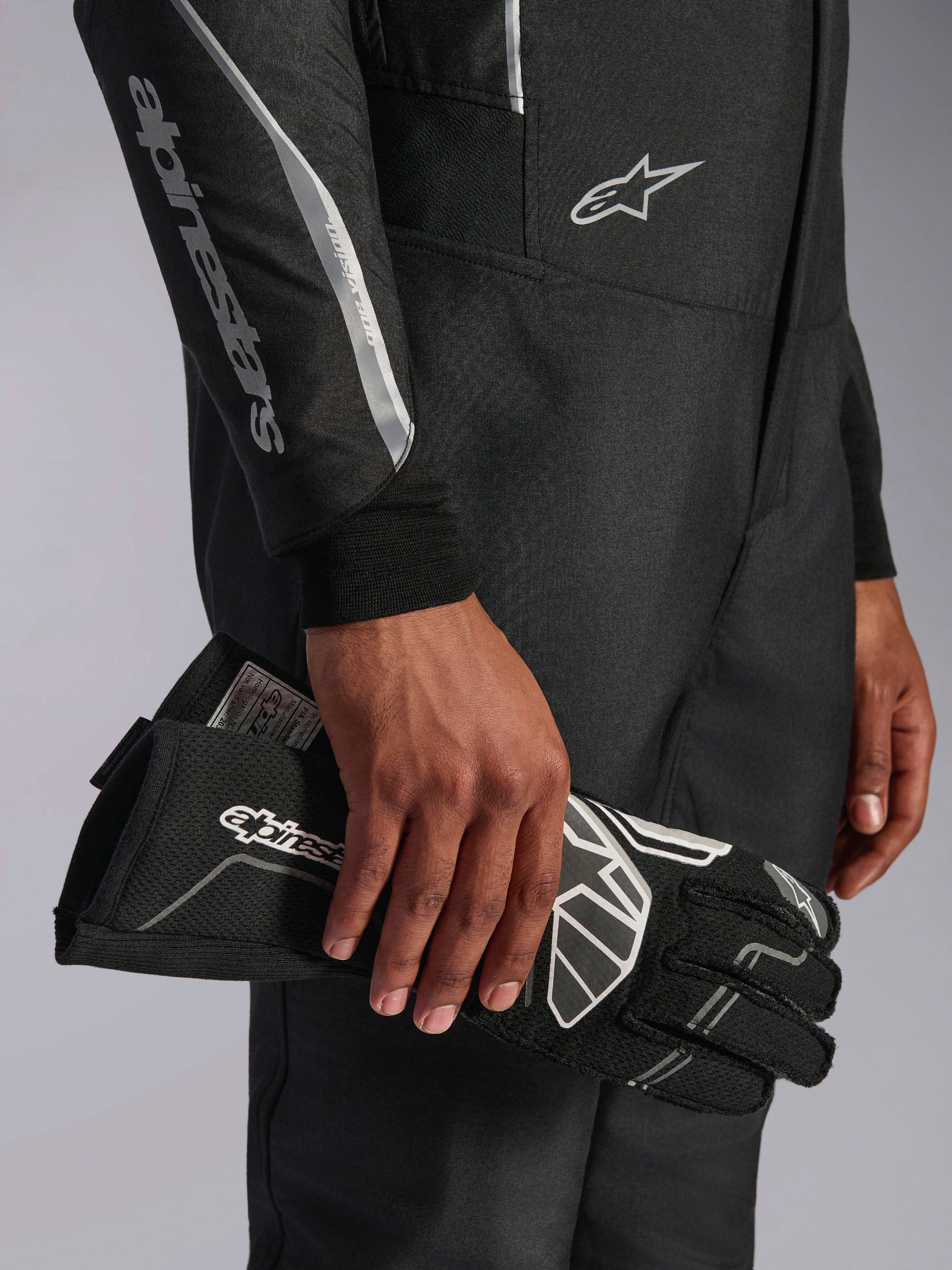 Alpinestars Hypertech V3 Suit in Black Dark Shadow, Nahaufnahme von Ärmel und Torso mit silbernem Alpinestars-Branding, Hand hält einen schwarzen Rennhandschuh mit weißen Akzenten, professionelle Autorennsport-Ausrüstung