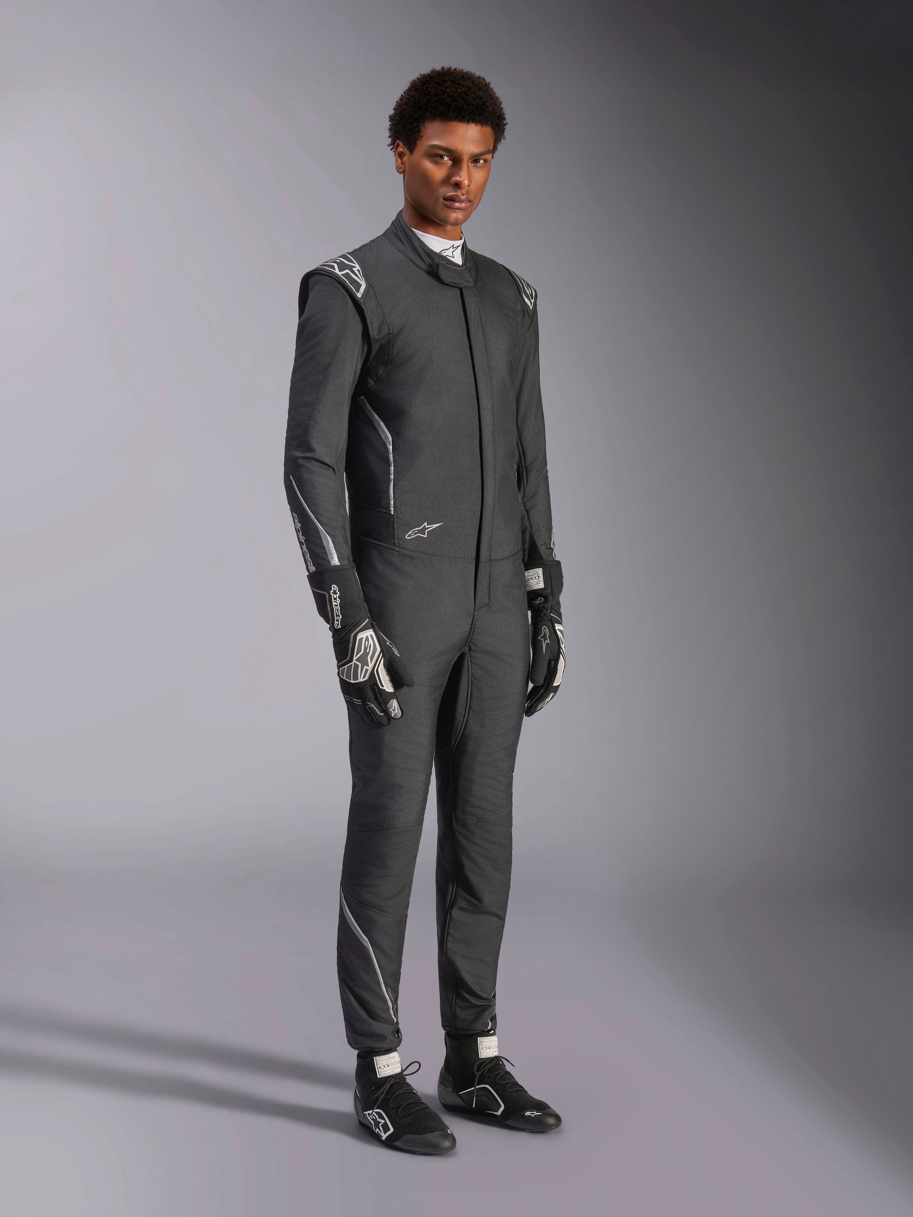 Alpinestars Hypertech V3 Anzug, Auto-Rennanzug, Schwarz und Dark Shadow, getragen von einem männlichen Model mit passenden schwarzen Handschuhen und Rennschuhen, sportlich schlanke Passform, mit Alpinestars-Logos und hellgrauen Akzenten, professionelle Motorsport-Schutzausrüstung