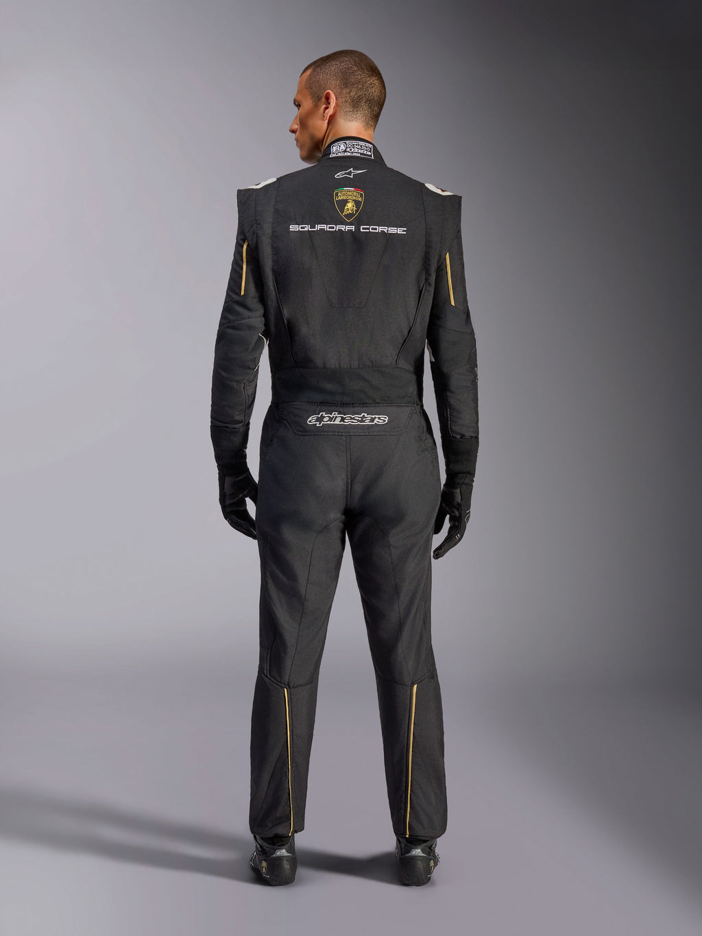 Alpinestars Limitierte Edition Lamborghini GP Tech V4 Anzug FIA, Automobil-Rennanzug, Schwarz mit gold- und silberfarbenen Akzenten, Rückansicht mit Lamborghini Squadra Corse Branding, Alpinestars-Logo und goldener Paspelierung an Ärmeln und Beinen, professionelle Rennausrüstung