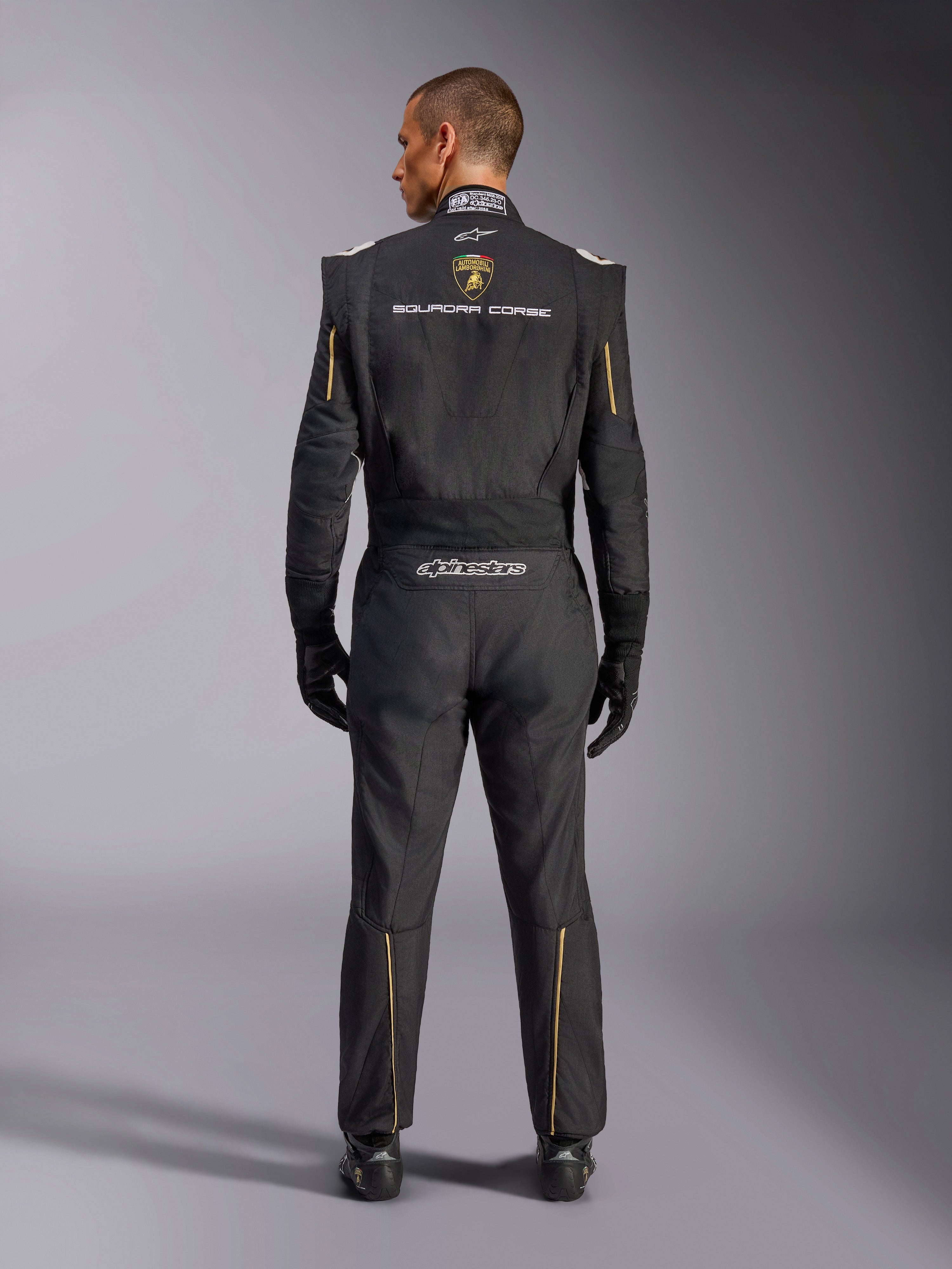 Alpinestars Limitierte Edition Lamborghini GP Tech V4 Anzug FIA, Automobil-Rennanzug, Schwarz mit gold- und silberfarbenen Akzenten, Rückansicht mit Lamborghini Squadra Corse Branding, Alpinestars-Logo und goldener Paspelierung an Ärmeln und Beinen, professionelle Rennausrüstung