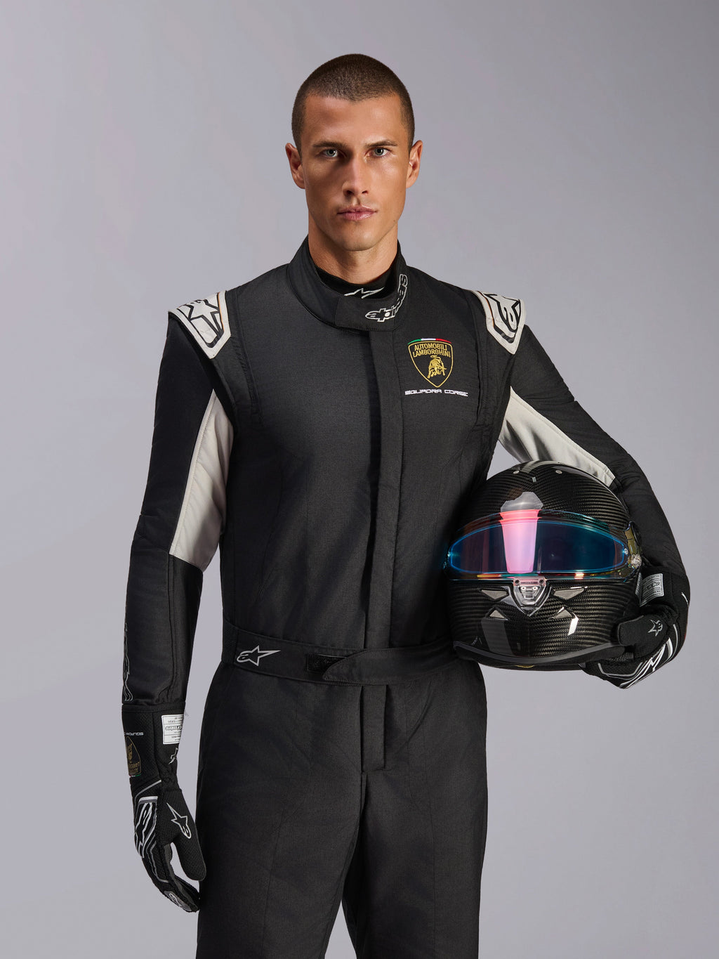 Alpinestars Limited Edition Lamborghini GP Tech V4 Anzug, Autorennanzug, Schwarz, Silber und Gold, getragen von einem Modell, das einen Carbon-Helm mit irisierendem Visier hält, mit Lamborghini Squadra Corse Branding und weißen Schulterakzenten