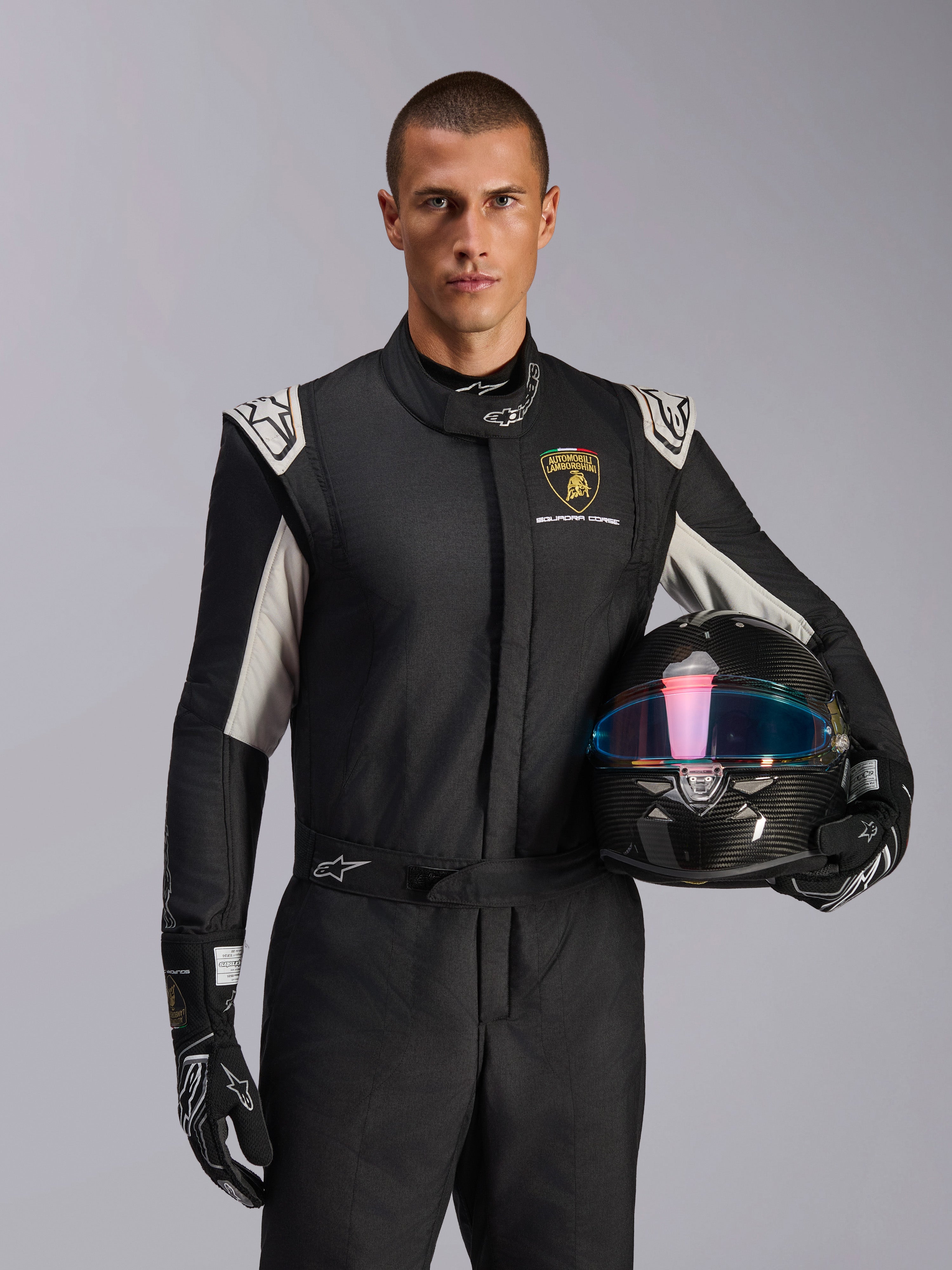 Alpinestars Limited Edition Lamborghini GP Tech V4 Anzug, Autorennanzug, Schwarz, Silber und Gold, getragen von einem Modell, das einen Carbon-Helm mit irisierendem Visier hält, mit Lamborghini Squadra Corse Branding und weißen Schulterakzenten
