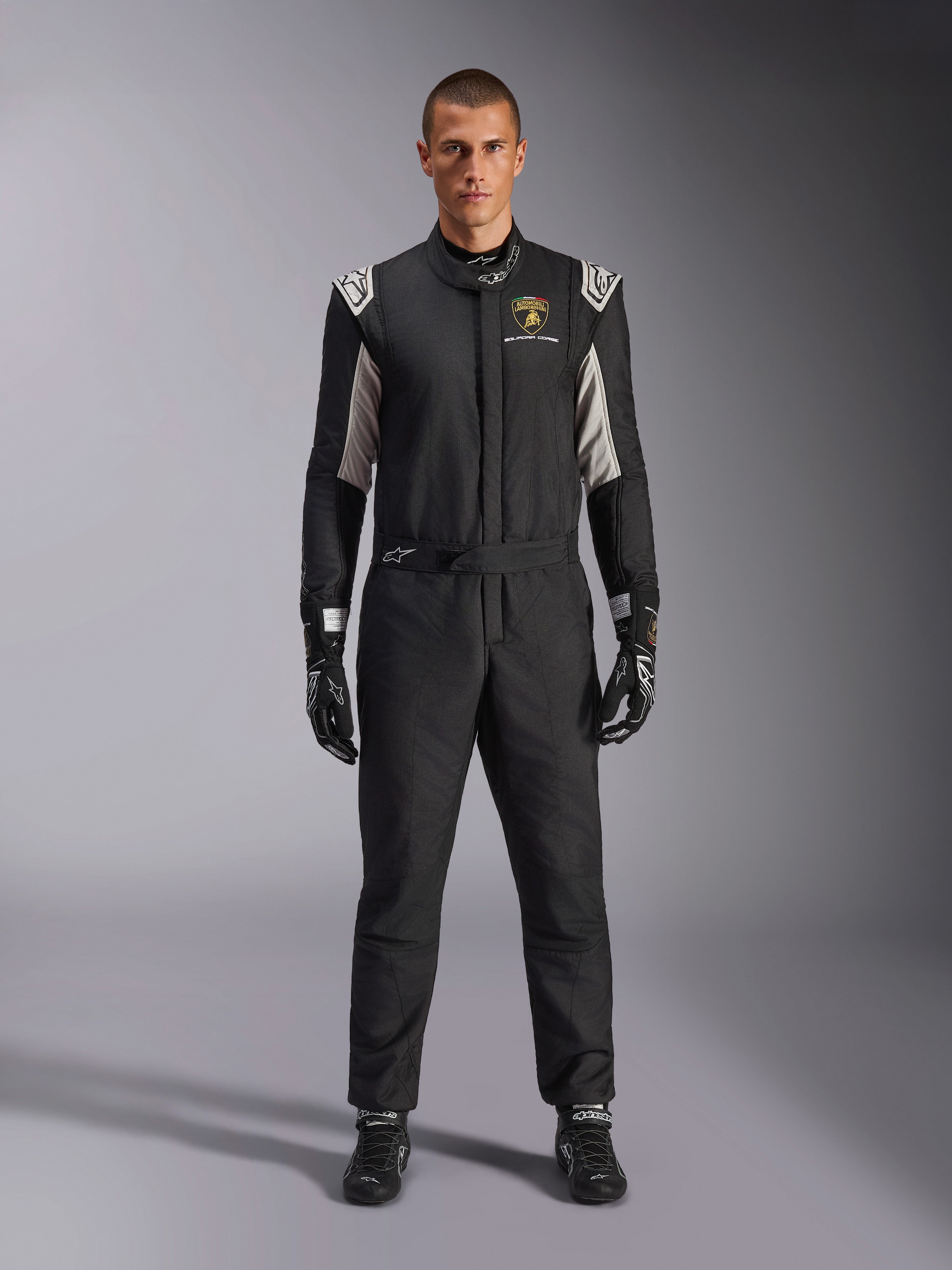 Alpinestars Limited Edition Lamborghini GP Tech V4 Anzug FIA, professioneller Autorennanzug, Schwarz mit silbernen und goldenen Akzenten, mit Automobili Lamborghini Squadra Corse Logo auf der Brust, getragen mit passenden Rennhandschuhen und Stiefeln, schlankes sportliches Design für Rundstreckenrennen