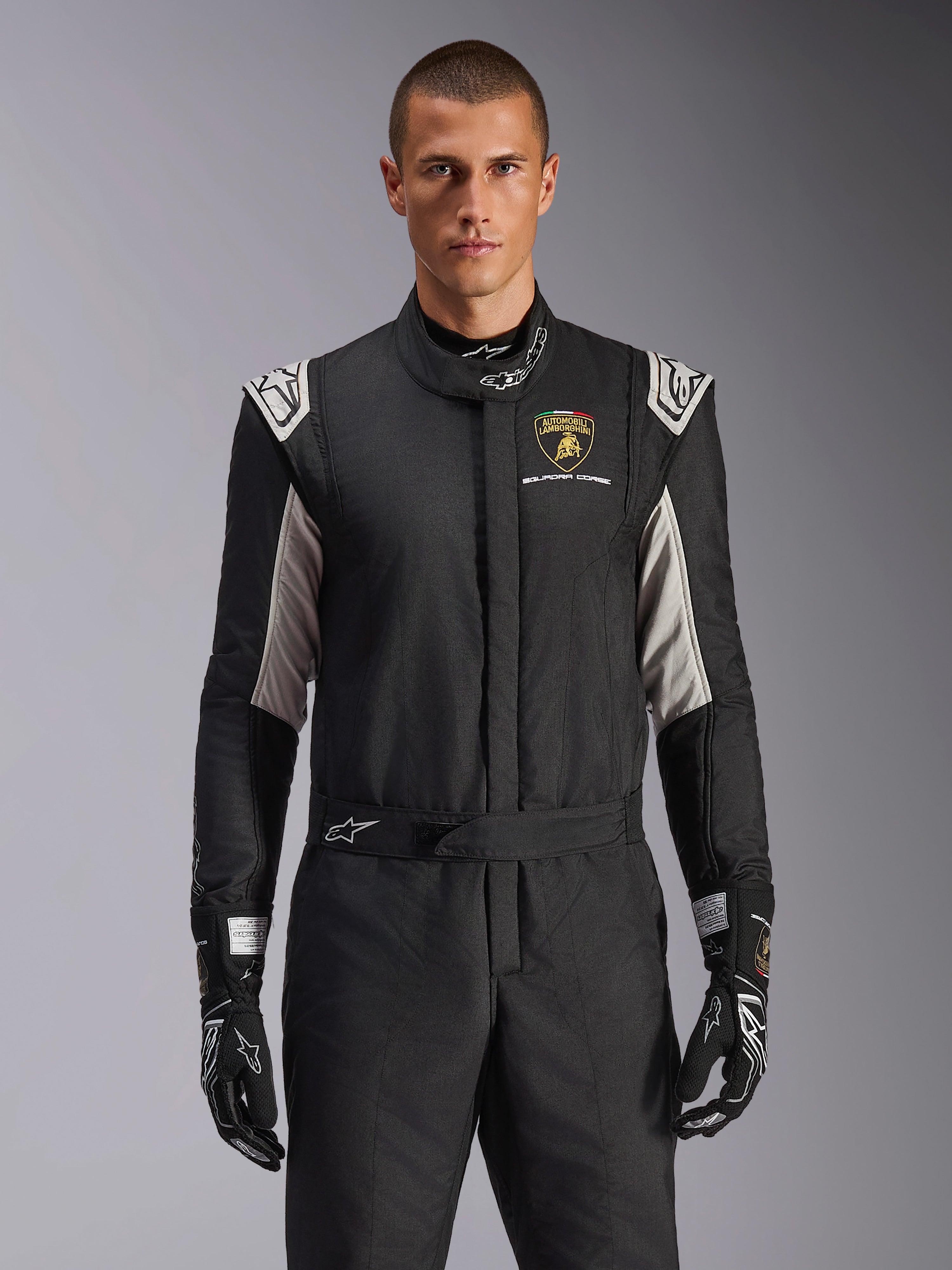 Alpinestars Limited Edition Lamborghini GP Tech V4 Anzug, professioneller Rennanzug in Schwarz mit silbernen und goldenen Akzenten, mit dem Automobili Lamborghini Squadra Corse Wappen auf der Brust, getragen von einem Model mit passenden schwarzen Rennhandschuhen
