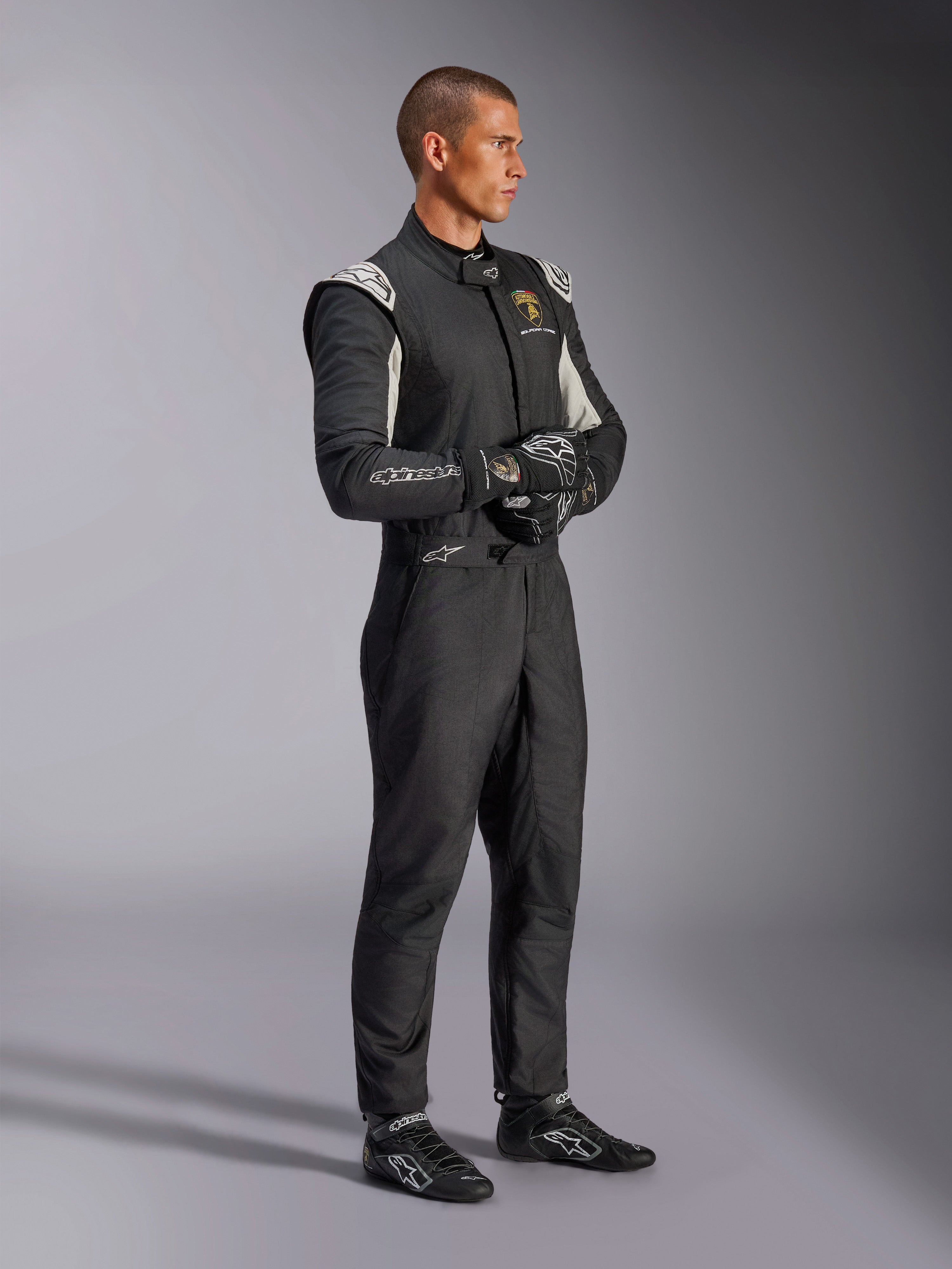 Männlicher Fahrer im Alpinestars Limited Edition Lamborghini GP Tech V4 Anzug, FIA-zertifizierter Autorennanzug in Schwarz, Silber und Gold, kombiniert mit passenden Rennhandschuhen und -schuhen, mit Lamborghini Squadra Corse Branding, professionelle Motorsport-Rennausrüstung