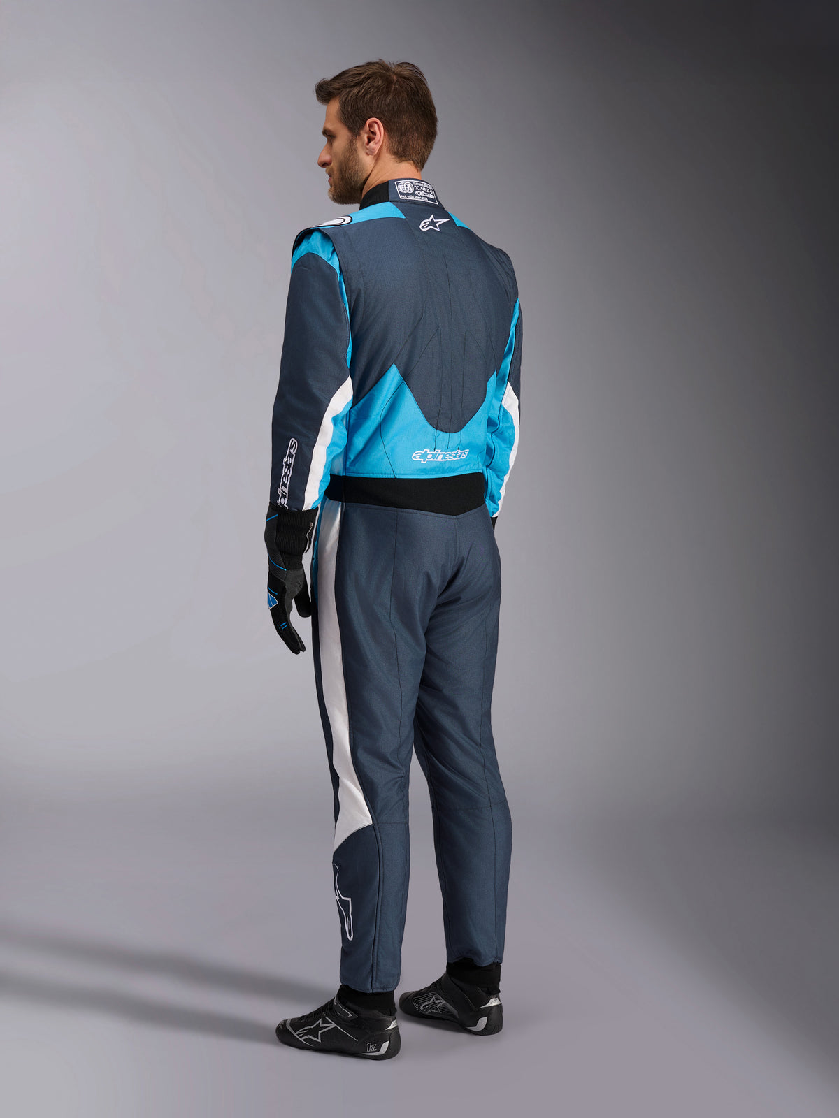 Alpinestars GP Pro Comp V2 FIA Auto Racing Suit in Asphalt and Cyan blue, rear view on a model with professional racing gear.