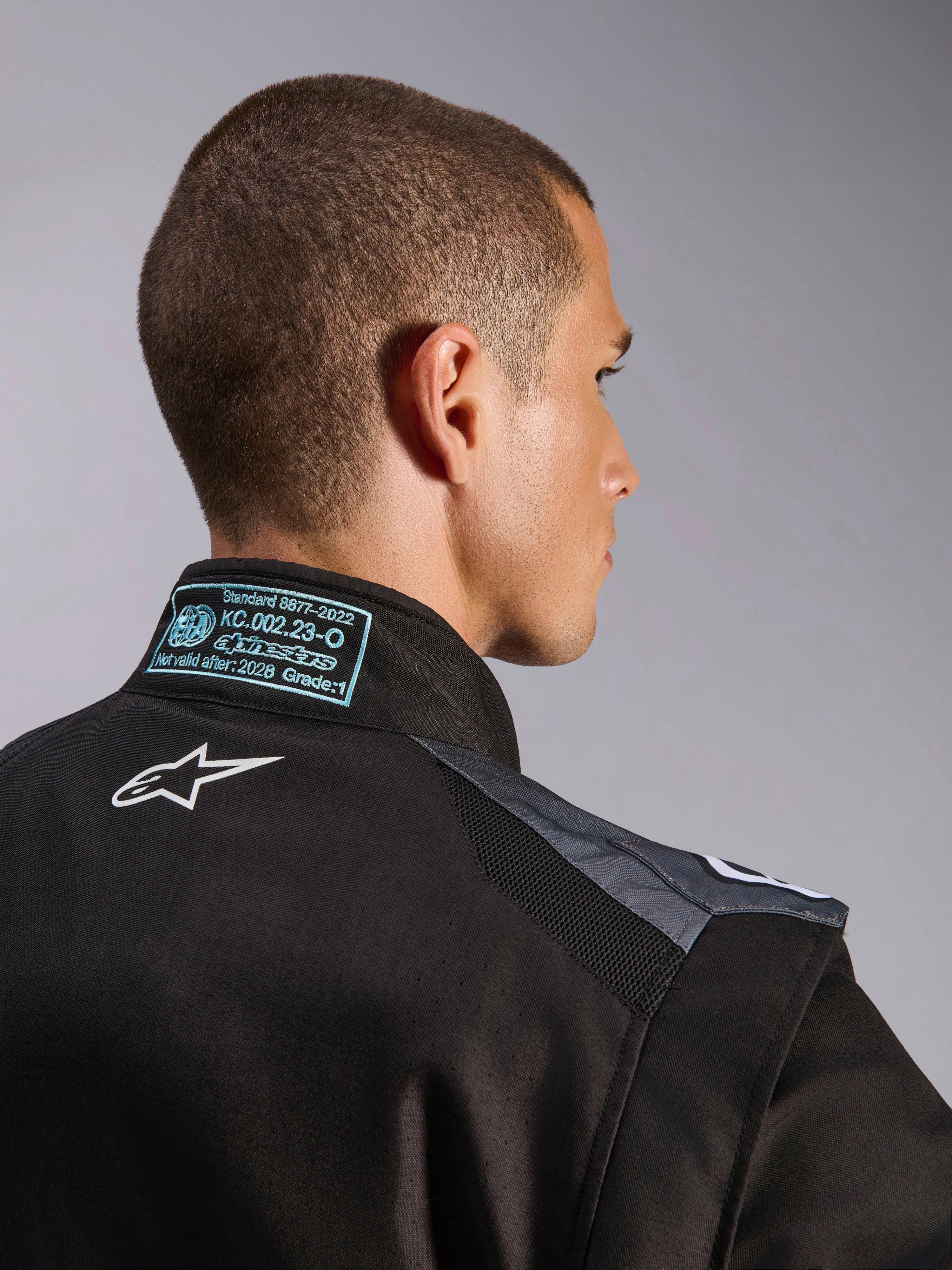 Alpinestars KMX-5 V3 Anzug in Schwarz und Dark Shadow, Nahaufnahme von Rücken und Kragen mit weißem Astars-Logo, FIA 8877-2022 Grade 1 Zertifizierungsetikett und belüfteten Mesh-Schultereinsätzen.