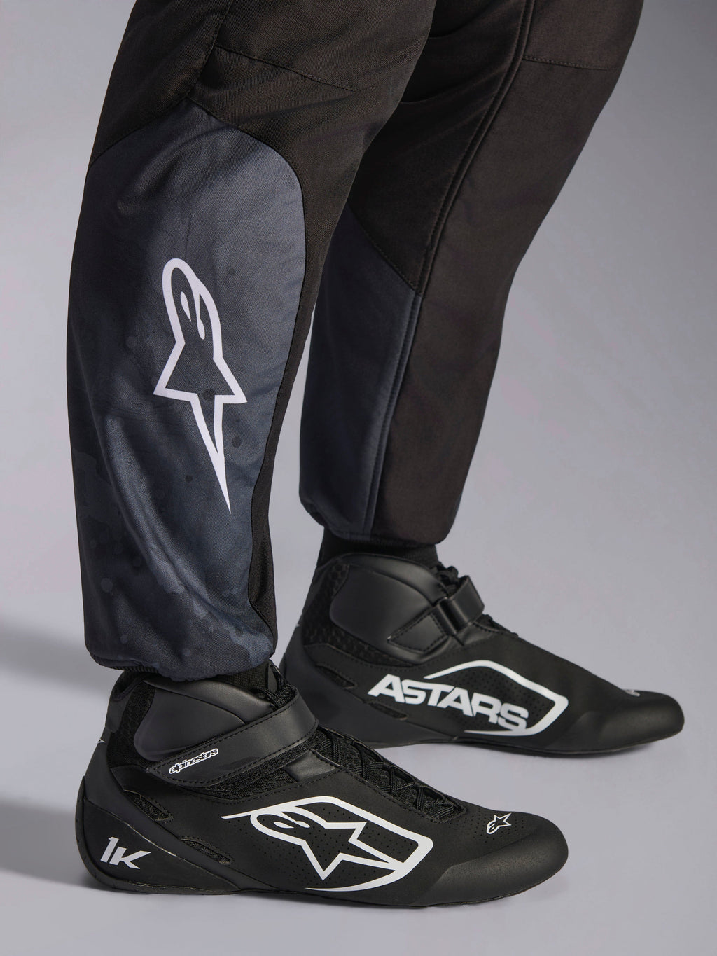 Alpinestars KMX-5 V3 Anzug, Schwarz Dark Shadow, Nahaufnahme der Unterschenkel und Knöchel, weißes Stern-Logo auf der Wade, kombiniert mit schwarzen Kartschuhen, hebt die gestrickten Knöchelbündchen und die athletisch-reguläre Passform hervor.