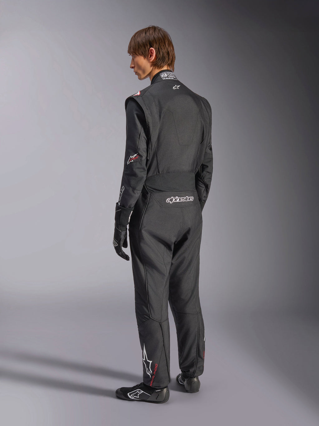 Alpinestars GP Tech V4 Anzug, Automobil-Rennanzug, Schwarz mit weißen Akzenten, Rückansicht eines Modells, das den Anzug mit Rennhandschuhen und Schuhen trägt, mit weißem Branding am unteren Rücken und am Bein, professionelle Motorsport-Schutzausrüstung