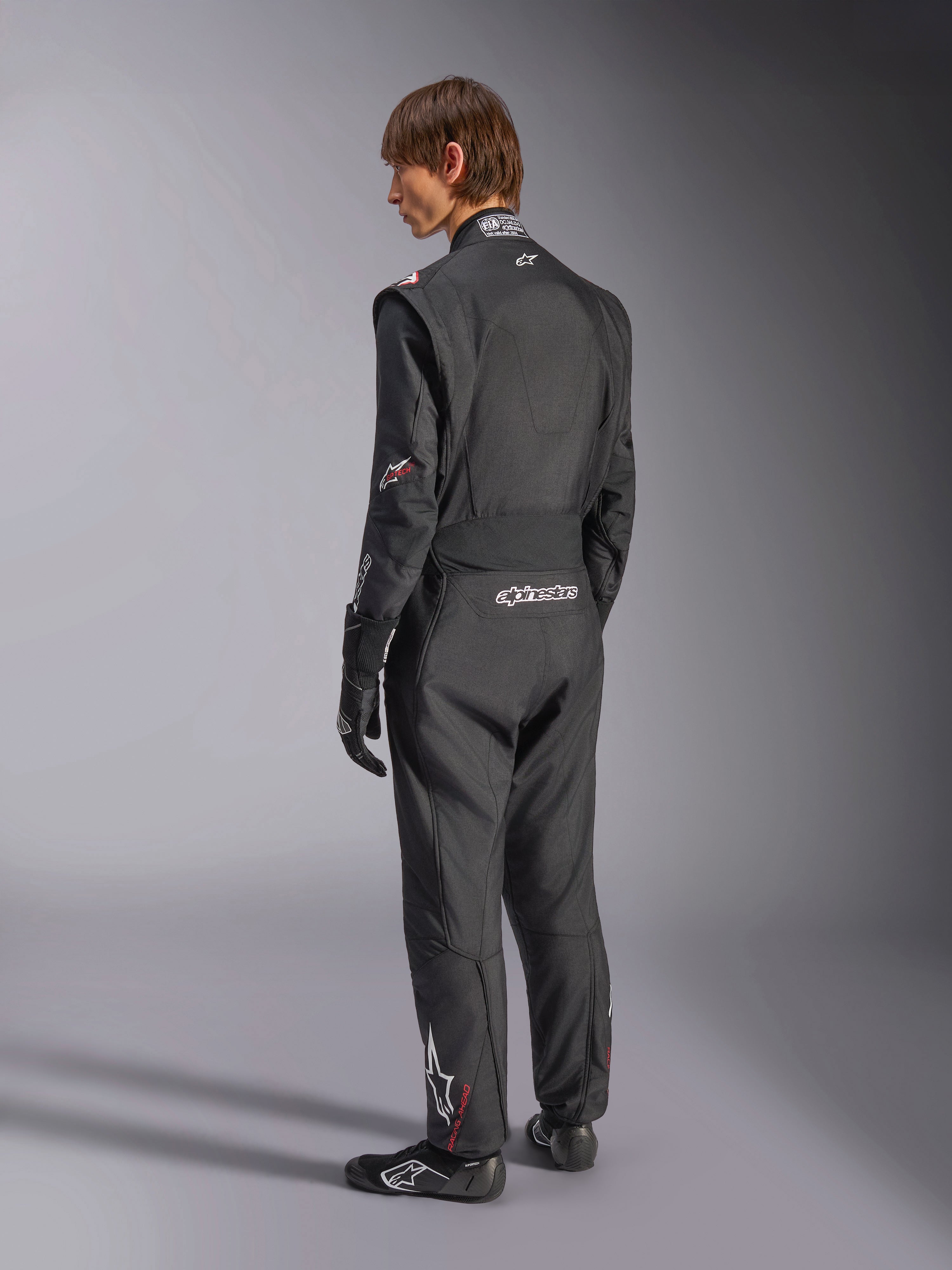 Alpinestars GP Tech V4 Anzug, Automobil-Rennanzug, Schwarz mit weißen Akzenten, Rückansicht eines Modells, das den Anzug mit Rennhandschuhen und Schuhen trägt, mit weißem Branding am unteren Rücken und am Bein, professionelle Motorsport-Schutzausrüstung