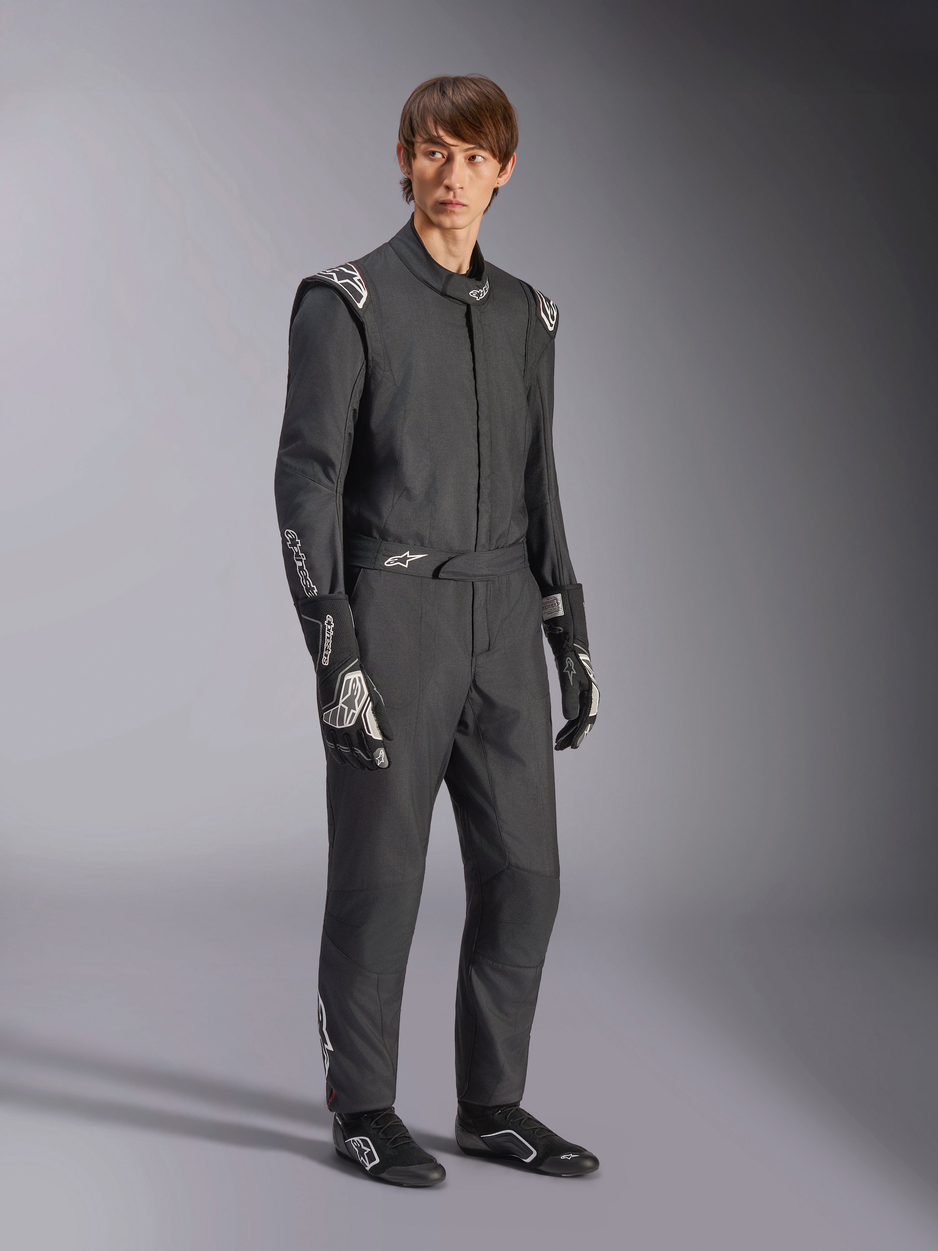 Alpinestars GP Tech V4 Anzug, einteiliger Autorennanzug, Schwarz und Weiß, getragen mit passenden Rennhandschuhen und Schuhen, mit weißen Logos auf den Schultern und Unterarmen, elegantes professionelles Design für den Motorsport