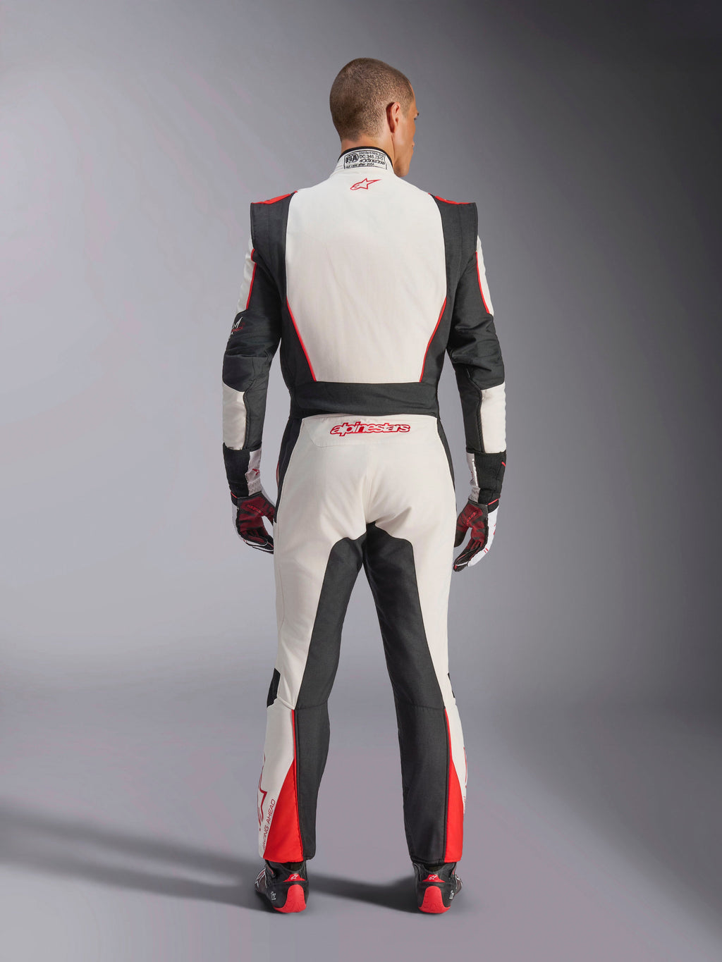 Alpinestars GP Tech V4 Anzug, Auto-Rennanzug, Weiß, Schwarz und Rot, Rückansicht eines Fahrers im einteiligen Anzug, mit schwarzen Stretcheinsätzen an den Seiten und Schultern, roten Akzenten an den Unterschenkeln, Alpinestars-Branding am unteren Rücken und am Kragen, professionelle FIA-zertifizierte Motorsportausrüstung