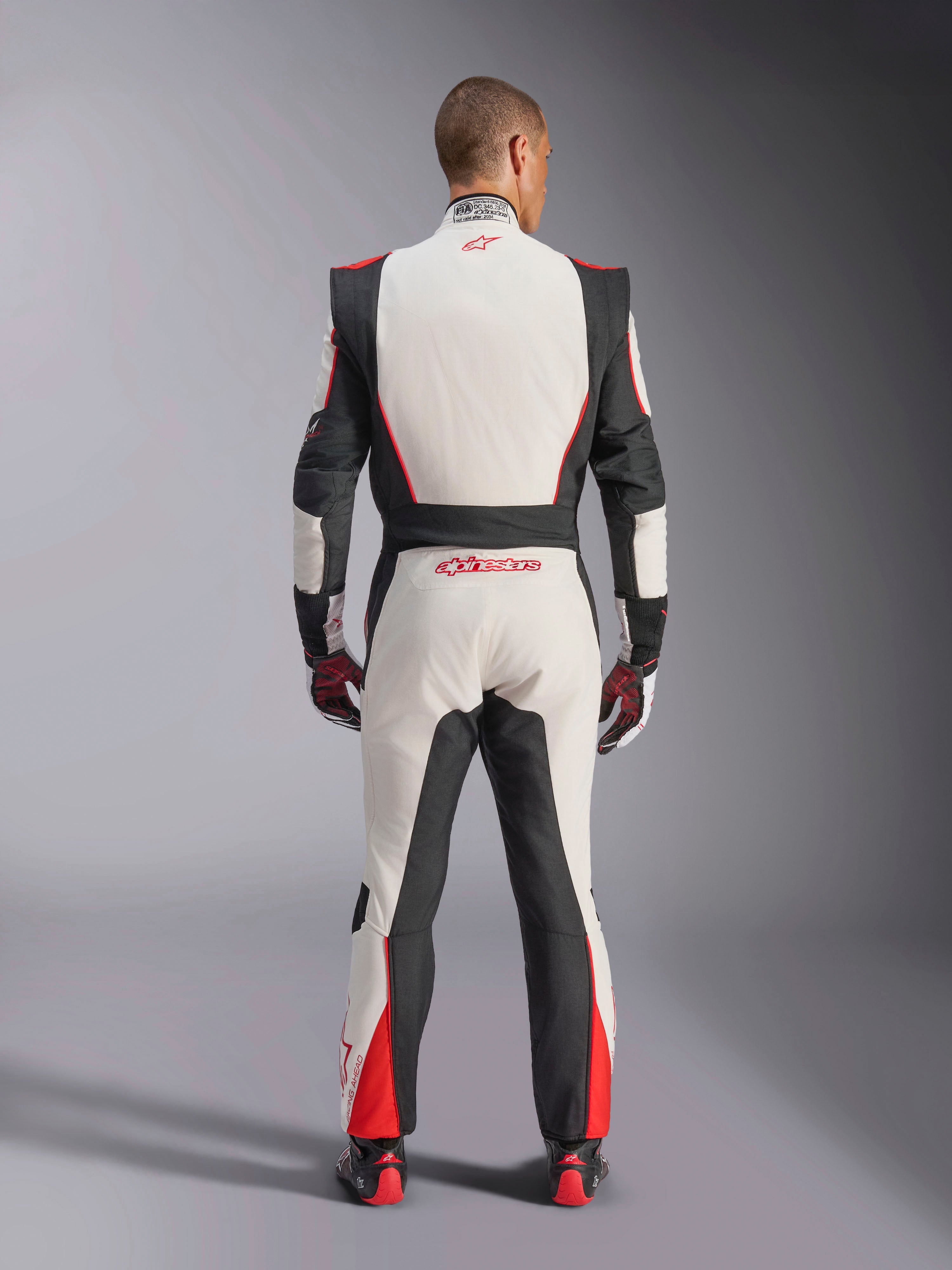 Alpinestars GP Tech V4 Anzug, Auto-Rennanzug, Weiß, Schwarz und Rot, Rückansicht eines Fahrers im einteiligen Anzug, mit schwarzen Stretcheinsätzen an den Seiten und Schultern, roten Akzenten an den Unterschenkeln, Alpinestars-Branding am unteren Rücken und am Kragen, professionelle FIA-zertifizierte Motorsportausrüstung