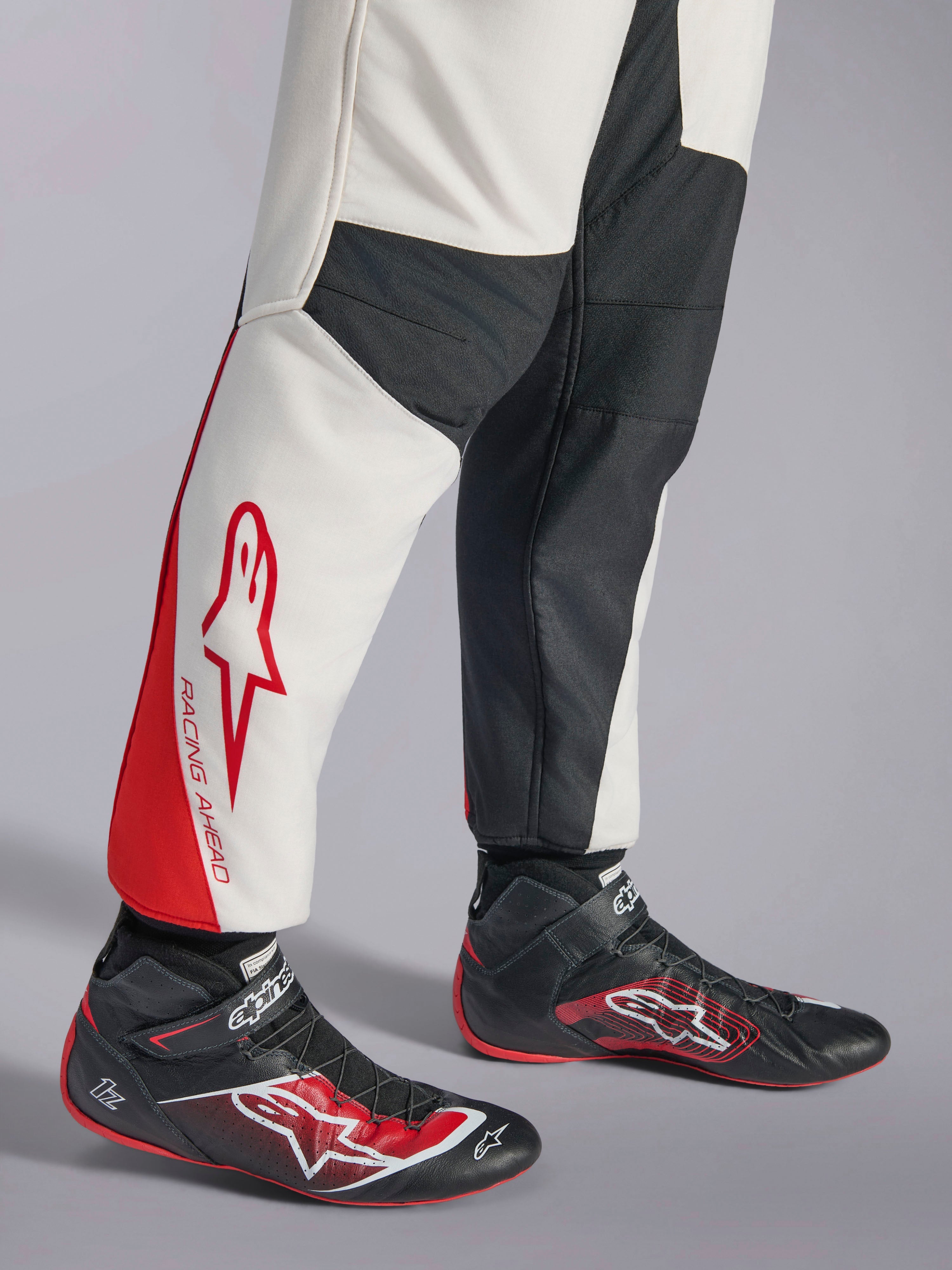 Alpinestars GP Tech V4 Anzug, Auto-Rennanzug, Weiß, Schwarz und Rot, Detailansicht der Unterschenkel mit weißen und schwarzen Paneelen, roten Akzenten und „RACING AHEAD“-Branding, getragen mit schwarzen Rennschuhen, professionelle Motorsportausrüstung