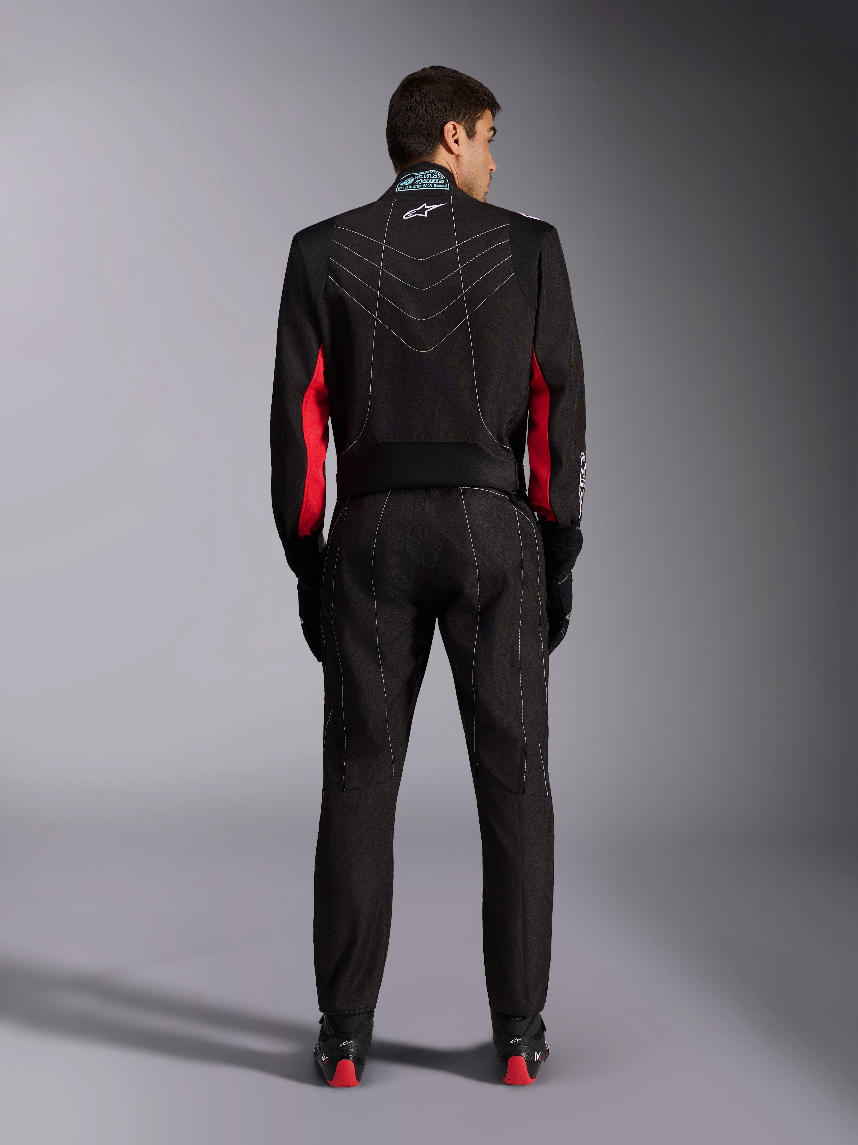Alpinestars KMX-9 V3 Anzug, Kart-Anzug, Schwarz mit roten und weißen Akzenten, Ganzkörperansicht von hinten, mit weißen V-förmigen Kontrastnähten, roten Einsätzen an den Ärmeln und Alpinestars-Logo am Kragen, kombiniert mit schwarzen Rennhandschuhen und Schuhen