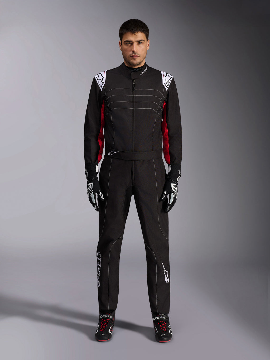 Alpinestars KMX-9 V3 Karting-Anzug in Schwarz Rot Weiß, Ganzkörper-Frontansicht eines Modells im einteiligen Anzug mit passenden Rennhandschuhen und Schuhen, mit weißen Alpinestars-Logos auf den Schultern und roten Akzenten.