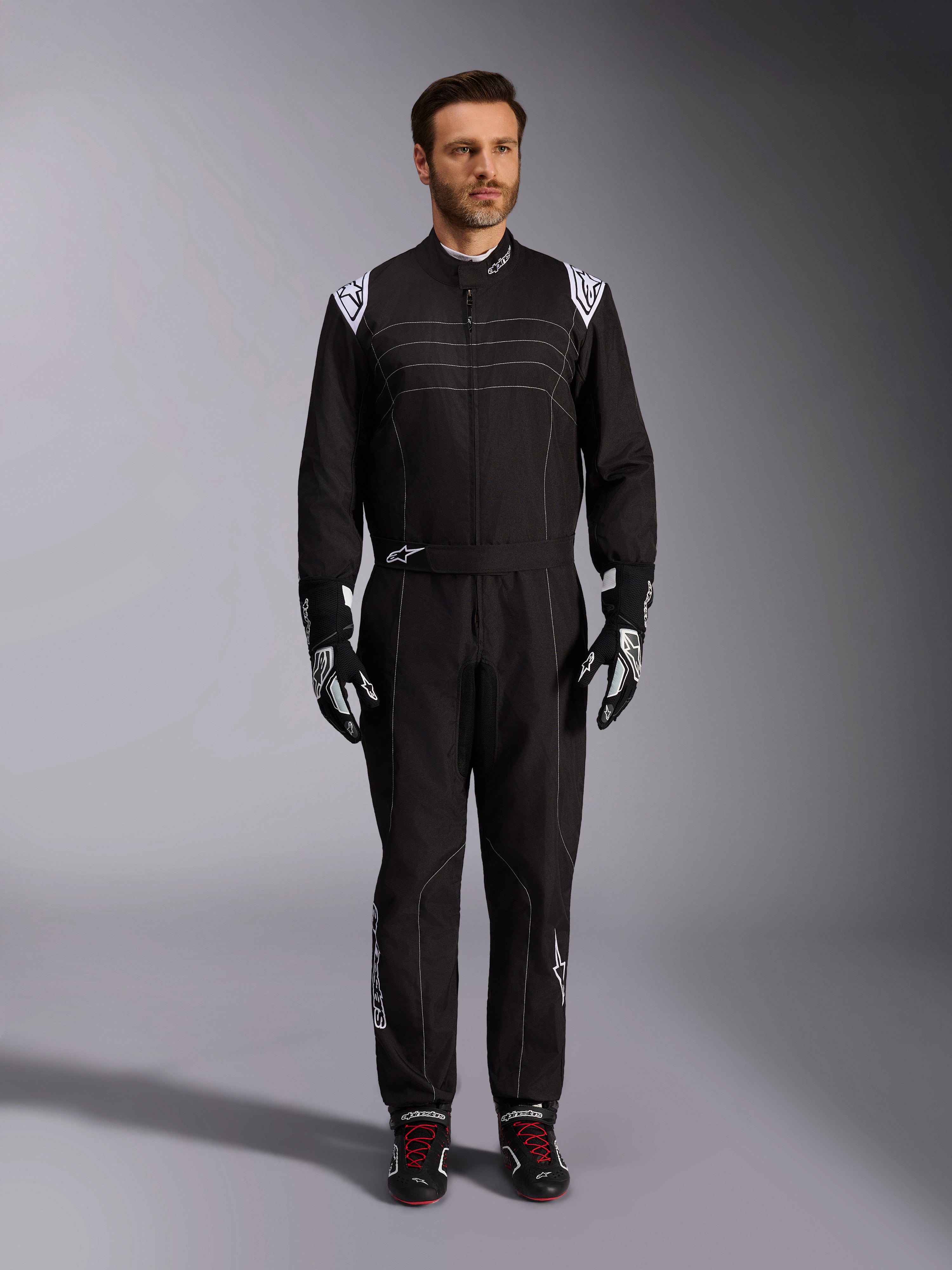 Alpinestars KMX-9 V3 Anzug, schwarz-weißer Kart-Anzug, Ganzkörperansicht an einem Modell, mit weißen Schulterlogos und Akzenten, kombiniert mit schwarz-weißen Rennhandschuhen und schwarzen Schuhen mit roten Schnürsenkeln, sportlich-reguläre Passform für professionelle Kart-Ausrüstung.