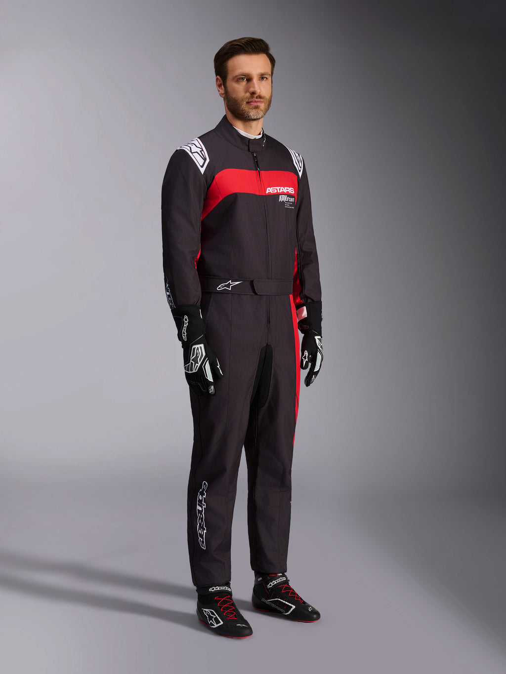 Männliches Modell im Alpinestars KMX-9 V3 Graphic 3 Anzug, Karting-Anzug, Schwarz und Rot, mit weißen Schulter-Epauletten und rotem Brust-Logo, kombiniert mit schwarzen Rennhandschuhen und Schuhen mit roten Schnürsenkeln, professionelle Motorsportbekleidung
