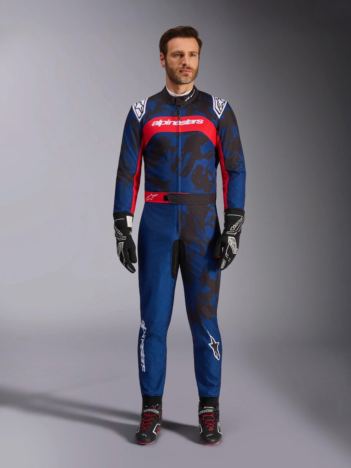 Alpinestars KMX-9 V3 Graphic 5 Anzug, Kart-Anzug in Navy-Blau und Rot, mit einem abstrakten grafischen Muster im Camouflage-Stil, getragen von einem Modell mit schwarzen Rennhandschuhen und Schuhen, sportliches Design mit markantem Alpinestars-Branding auf der Brust und den Beinen.