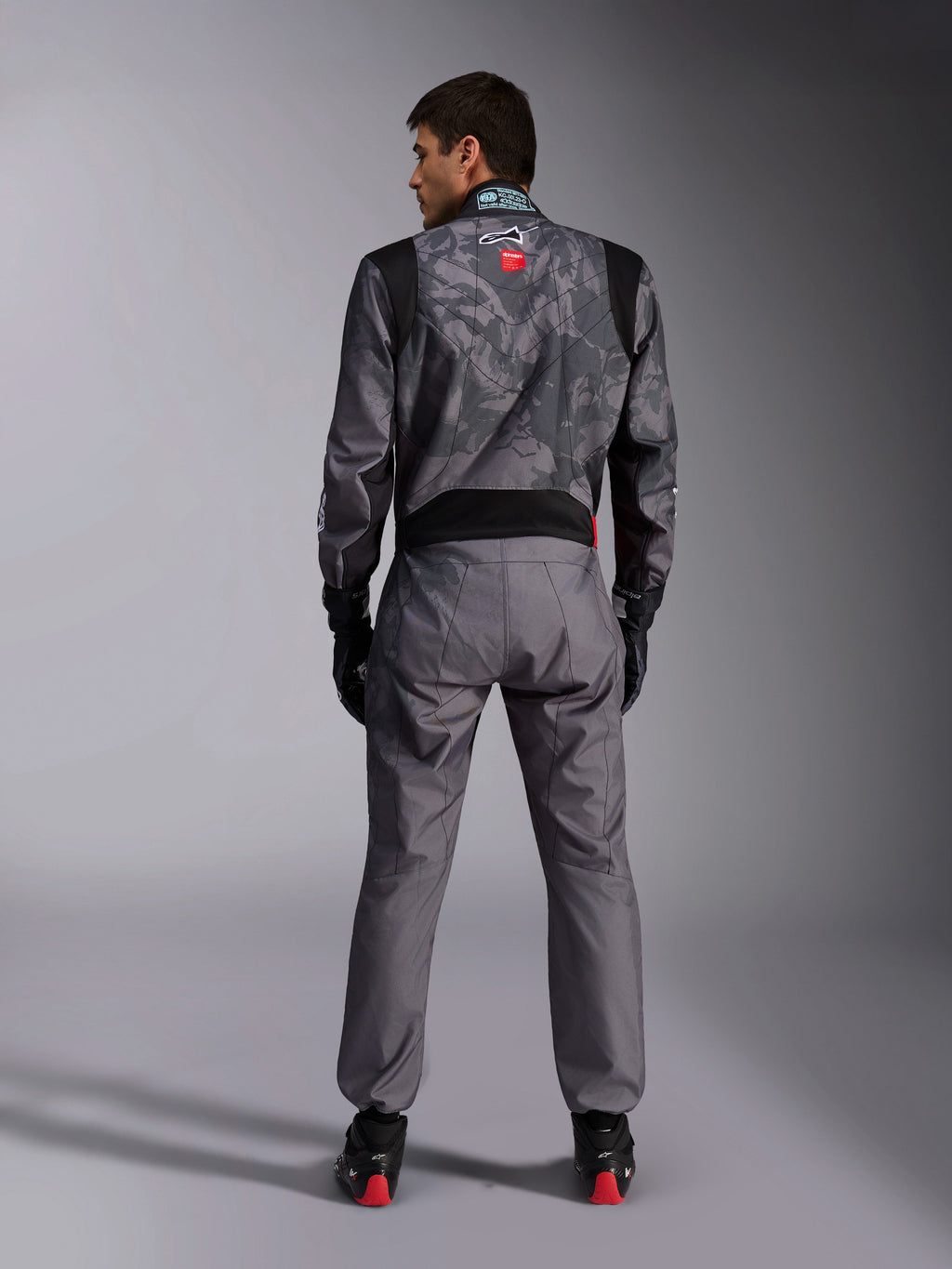 Alpinestars KMX-9 V3 Graphic 5 Anzug, Karting-Anzug, dunkelgraues und schwarzes Camouflage-Muster am Oberkörper, Rückansicht mit Stretcheinsätzen und Alpinestars-Branding, kombiniert mit schwarzen Handschuhen und schwarzen Rennschuhen mit roten Details, professionelle Motorsport-Rennausrüstung