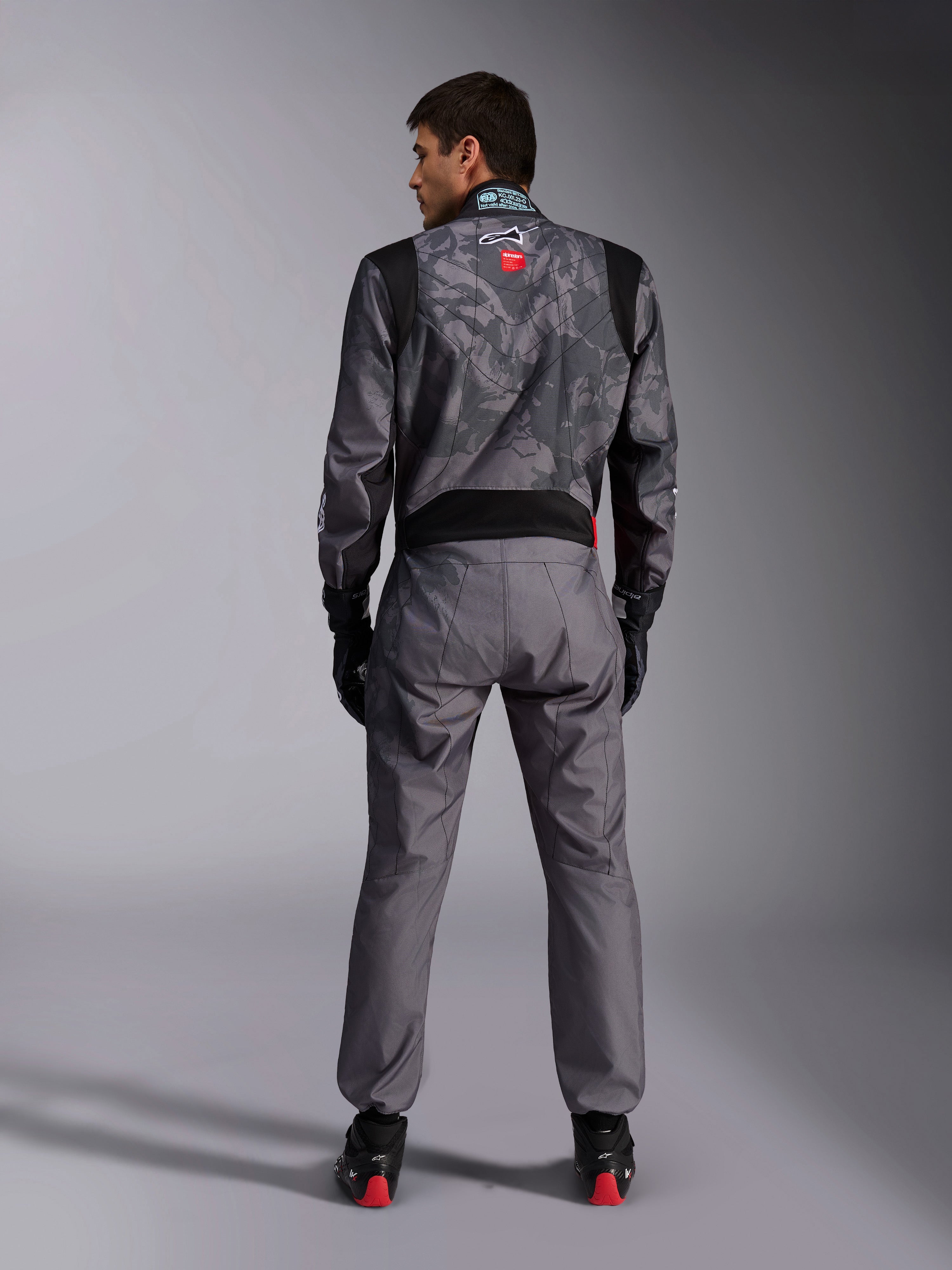 Alpinestars KMX-9 V3 Graphic 5 Anzug, Karting-Anzug, dunkelgraues und schwarzes Camouflage-Muster am Oberkörper, Rückansicht mit Stretcheinsätzen und Alpinestars-Branding, kombiniert mit schwarzen Handschuhen und schwarzen Rennschuhen mit roten Details, professionelle Motorsport-Rennausrüstung