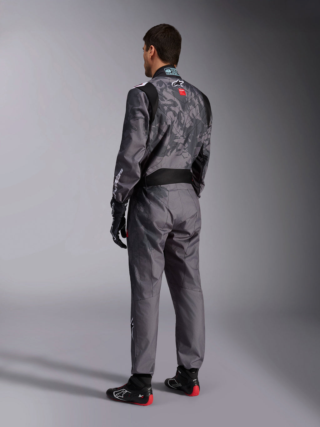 Alpinestars KMX-9 V3 Graphic 5 Anzug, dunkelgrauer und schwarzer Kart-Anzug, Rückansicht eines Models in kompletter Rennausrüstung einschließlich schwarz-roter Handschuhe und Kart-Schuhe, mit integrierten grafischen Mustern und Branding auf dem Rücken.