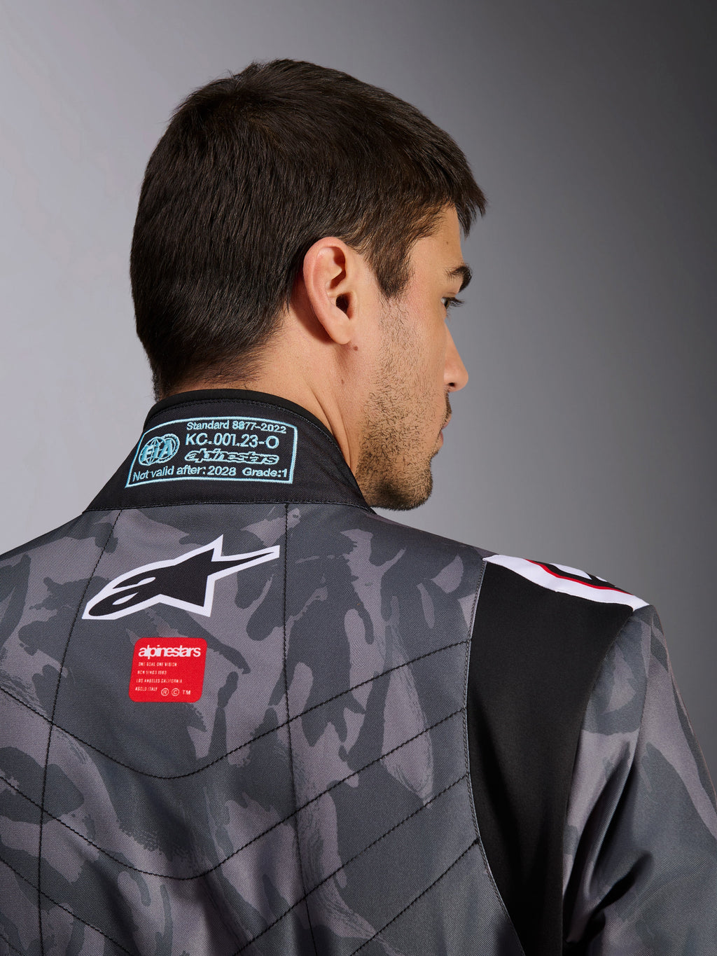 Alpinestars KMX-9 V3 Graphic 5 Anzug, Kart-Anzug, dunkelgraues und schwarzes Camouflage-Muster, Nahaufnahme des oberen Rückens mit Alpinestars-Logo, rotem Marken-Etikett und FIA-Homologationslabel am Kragen.