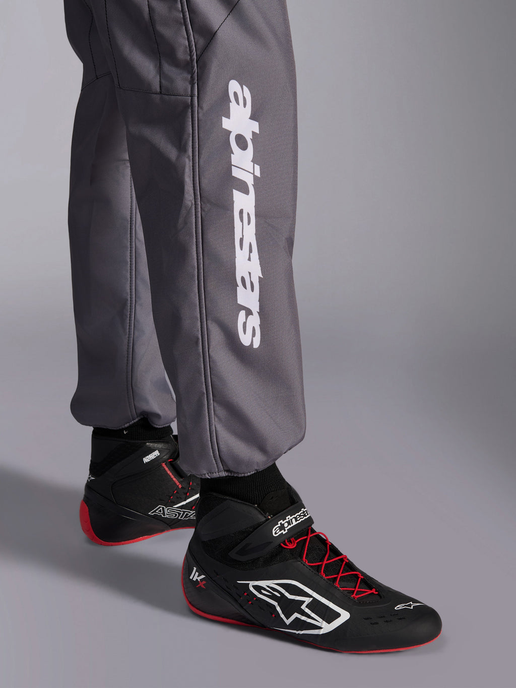 Alpinestars KMX-9 V3 Graphic 5 Anzug, Kart-Anzug, Dunkelgrau, Nahaufnahme des Unterschenkels mit weißem vertikalem Alpinestars-Logo, getragen mit schwarz-roten Rennschuhen, strapazierfähiges Material für den Kart-Wettbewerb