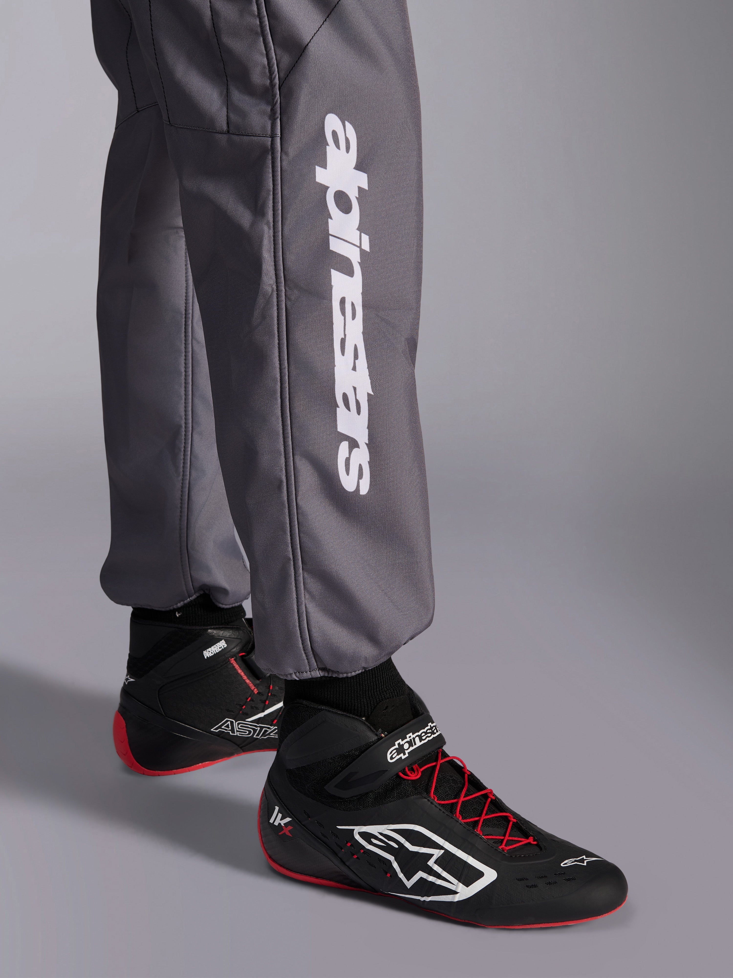 Alpinestars KMX-9 V3 Graphic 5 Anzug, Kart-Anzug, Dunkelgrau, Nahaufnahme des Unterschenkels mit weißem vertikalem Alpinestars-Logo, getragen mit schwarz-roten Rennschuhen, strapazierfähiges Material für den Kart-Wettbewerb