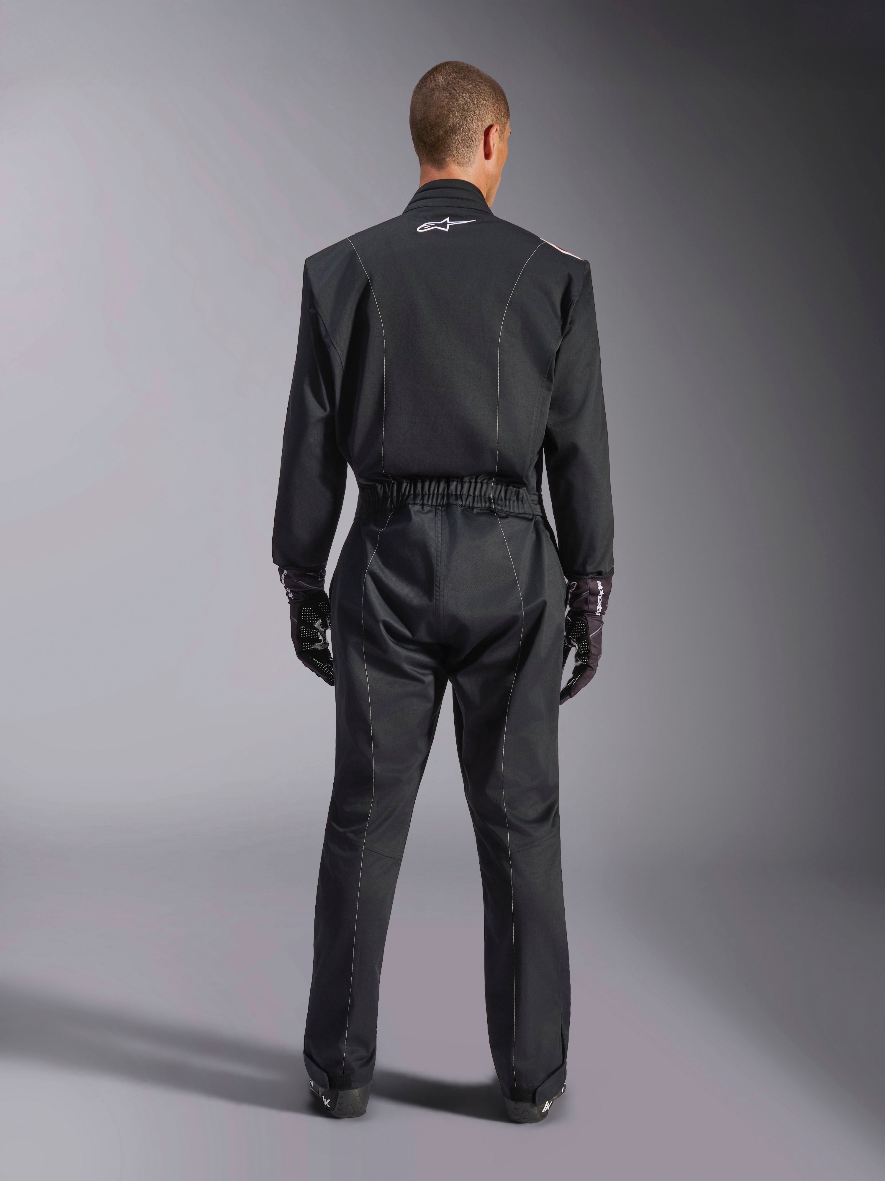 Alpinestars Kart Indoor V2 Suit, Black Karting Suit, rear full-body view on model, featuring white piping details and a small white Astars logo on the back collar, elastic waist belt, worn with black racing gloves, one-piece motorsport apparel.