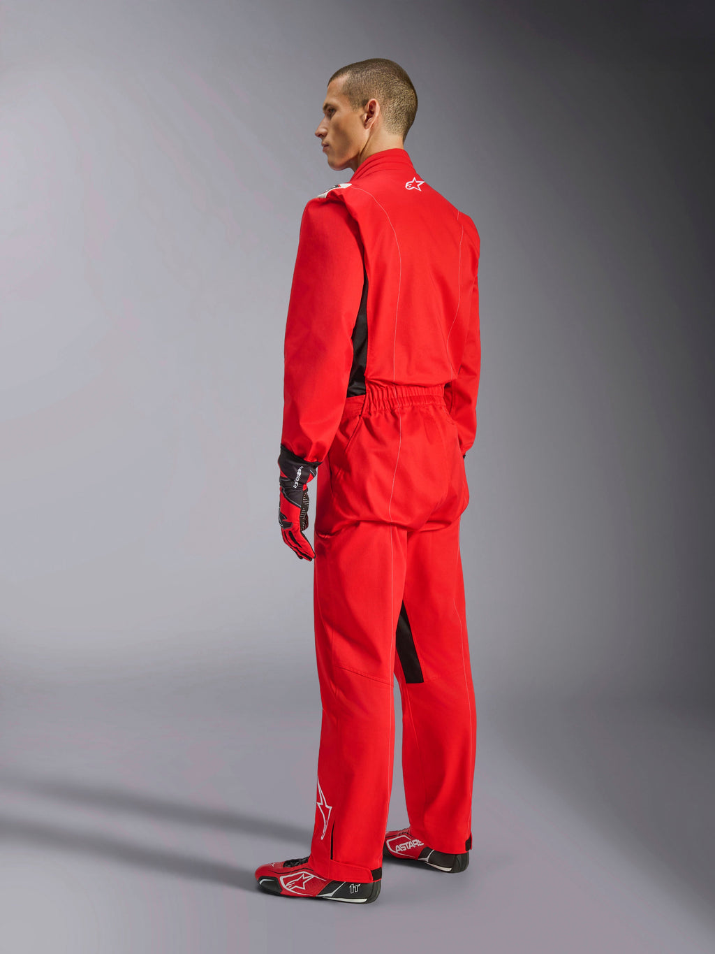 Alpinestars Kart Indoor V2 Anzug, roter Kart-Anzug, Dreiviertel-Rückansicht an einem Modell, getragen mit passenden roten Handschuhen und Rennschuhen, einlagige Poly-Baumwoll-Twill-Konstruktion, professionelle Motorsportausrüstung für Indoor-Kartfahren.