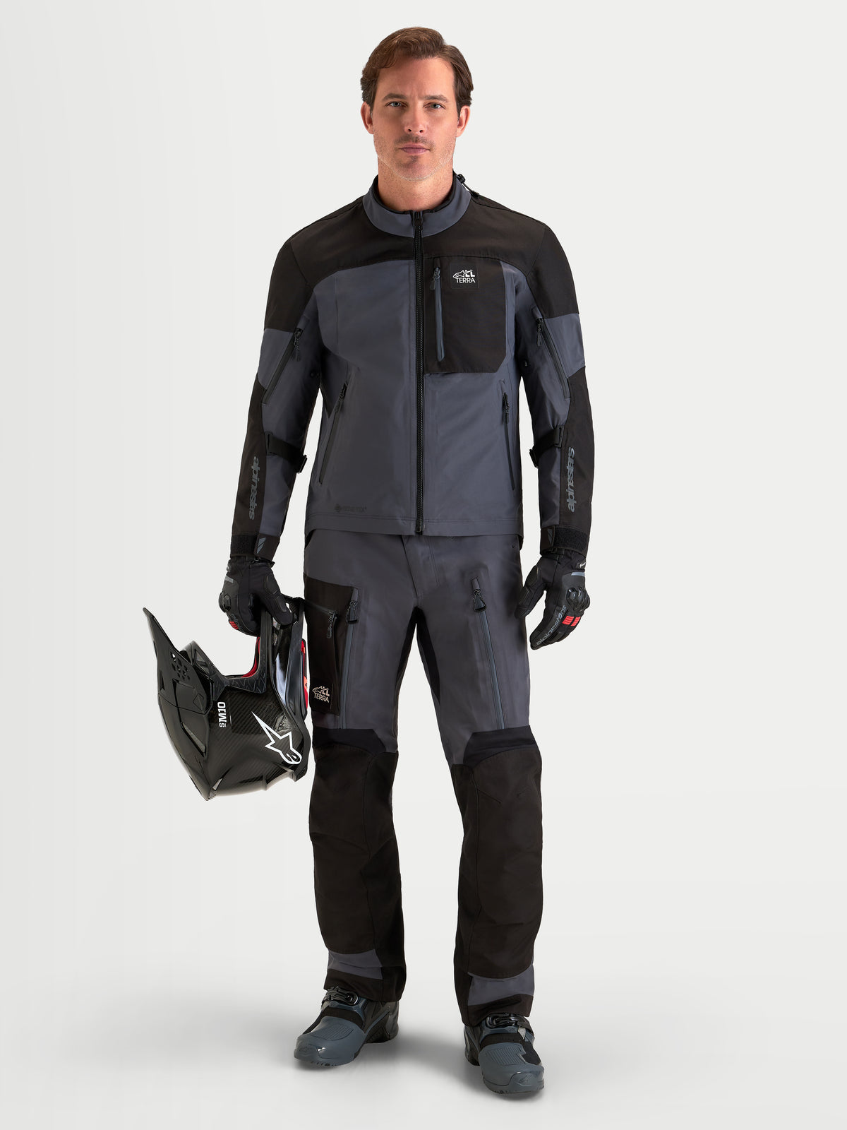 Alpinestars Arenis Gore-Tex Gear Set, Herren-Abenteuer-Motorradjacke und -hose in Schwarz und Dunkelgrau, Vorderansicht eines Models beim Tragen passender wasserdichter Textilausrüstung und Halten eines Helms