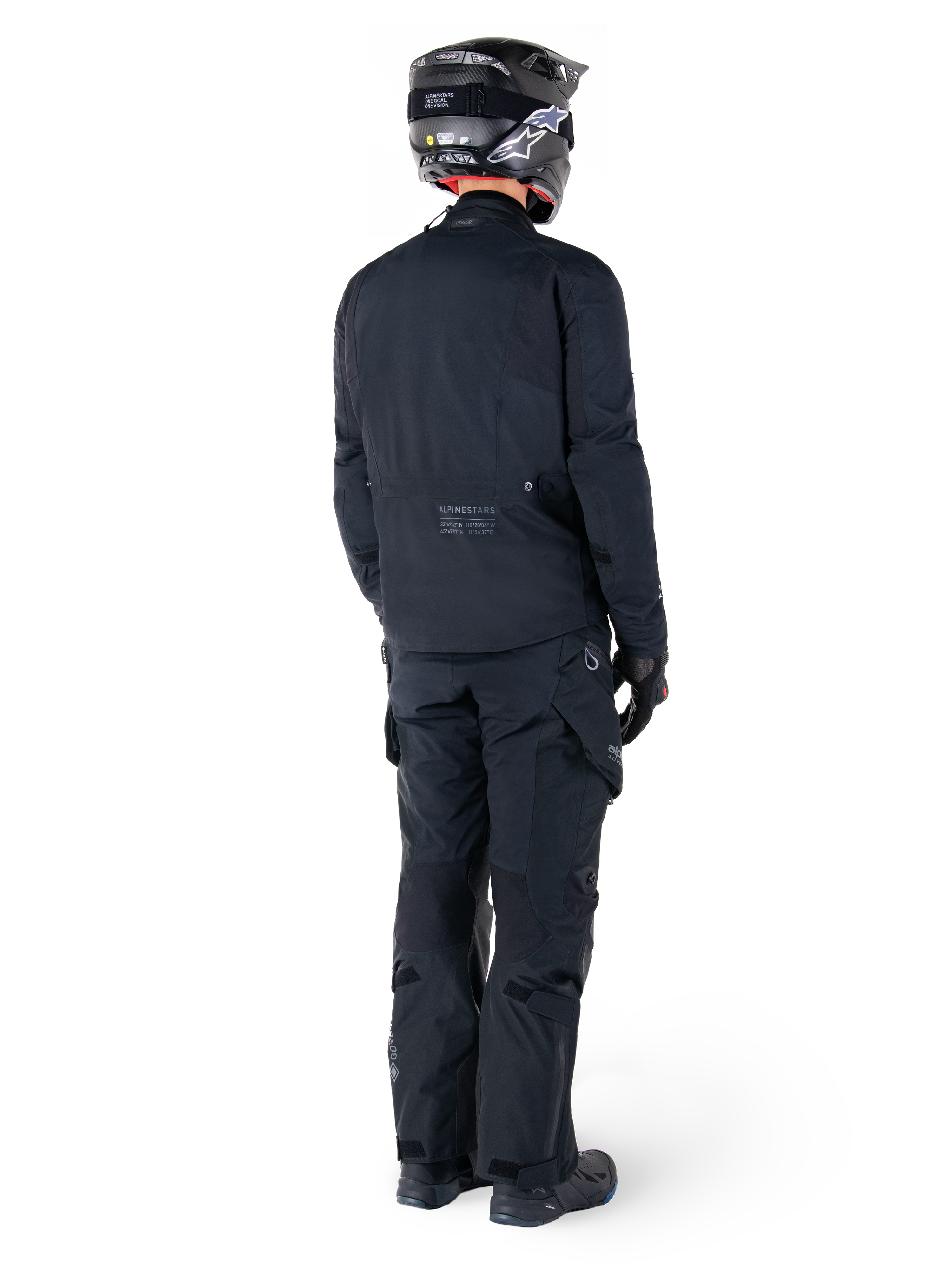 Rear view of a rider wearing the Alpinestars St-7 Prime GORE-TEX Jacket in black, paired with matching black touring pants, a black motorcycle helmet with white logo, and black riding boots, full protective adventure gear for touring and commuting.